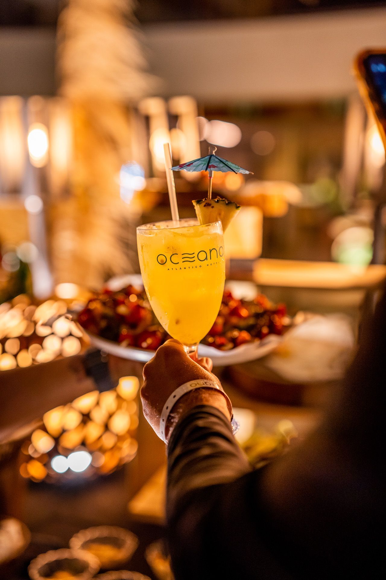 Pessoa segurando um coquetel decorado com uma fatia de abacaxi e um pequeno guarda-chuvinha; prato de aperitivos ao fundo.