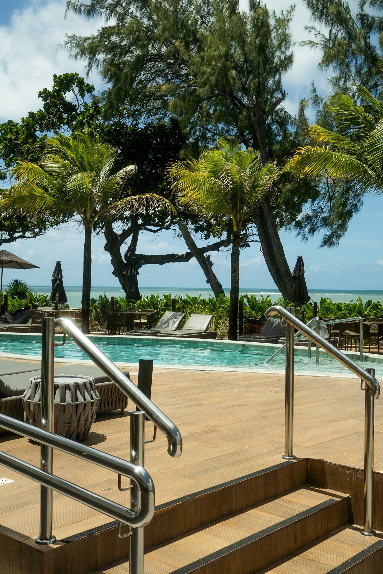 Área da piscina com escadas, corrimãos, árvores e vista para o mar em um dia ensolarado.