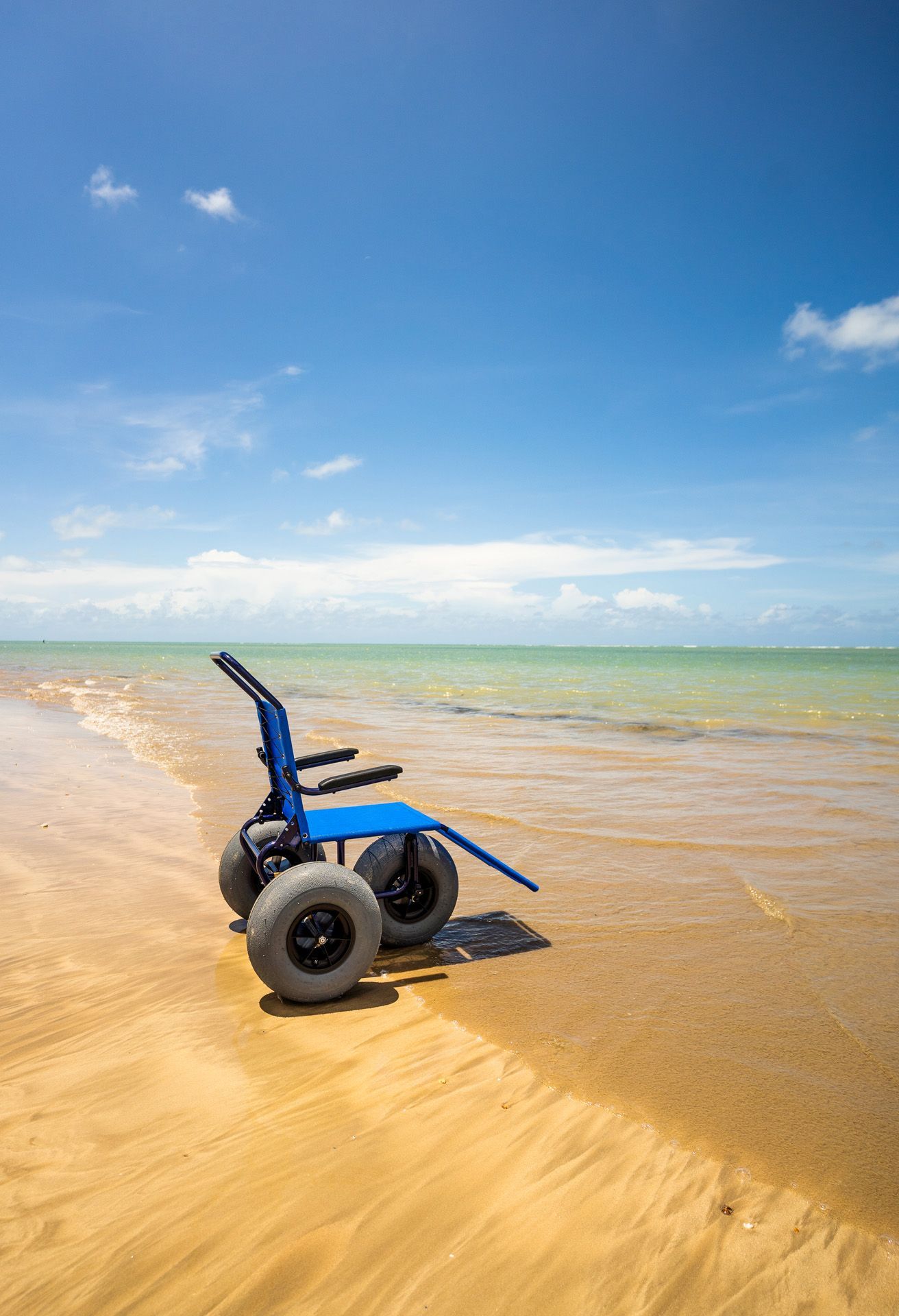 Caiaque azul e remos em uma praia de areia sob guarda-sóis, com o oceano e o céu azul ao fundo.