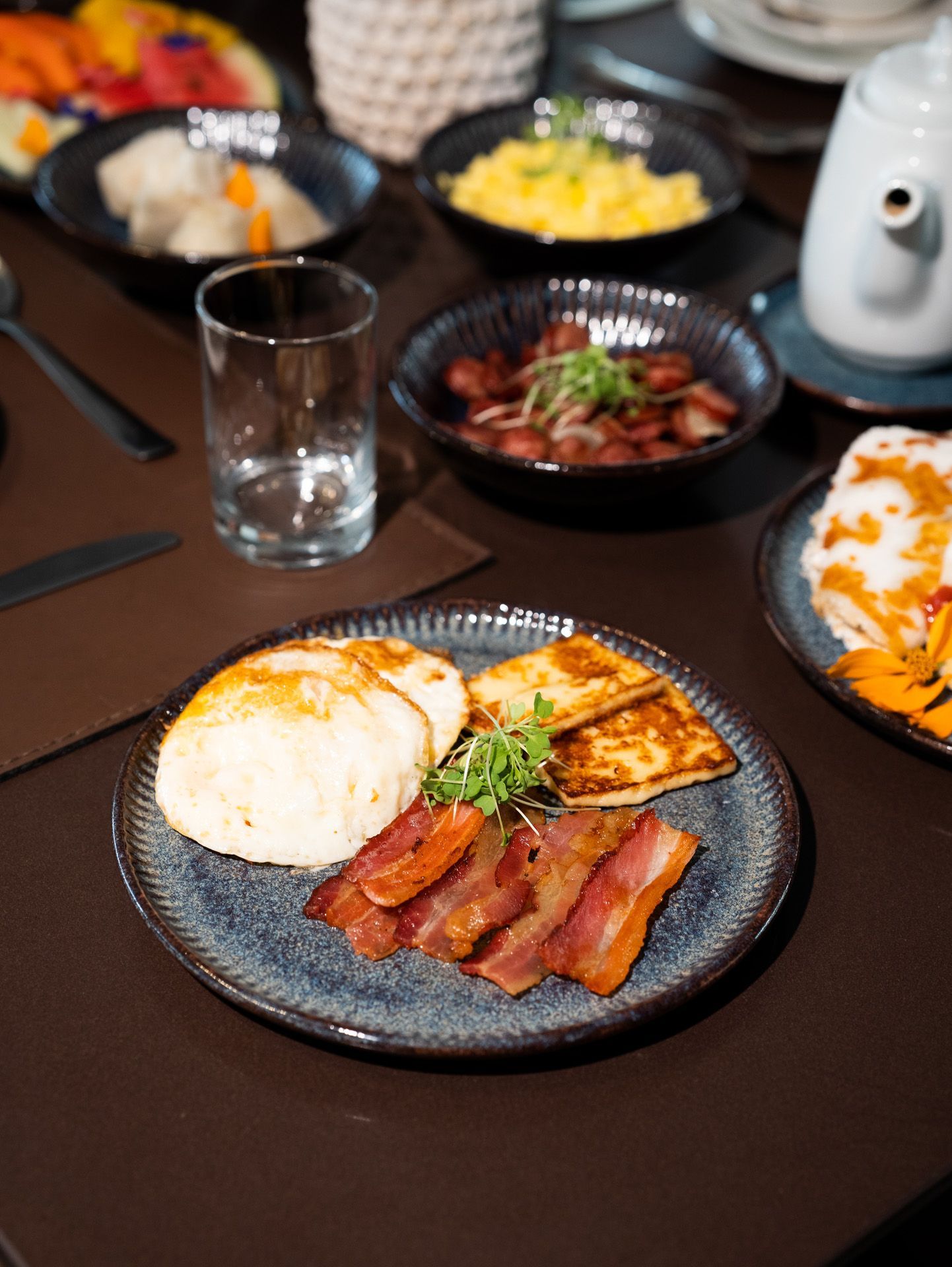 Mesa posta para o café da manhã: prato com bacon, pão e queijo, rodeado por outros pratos.