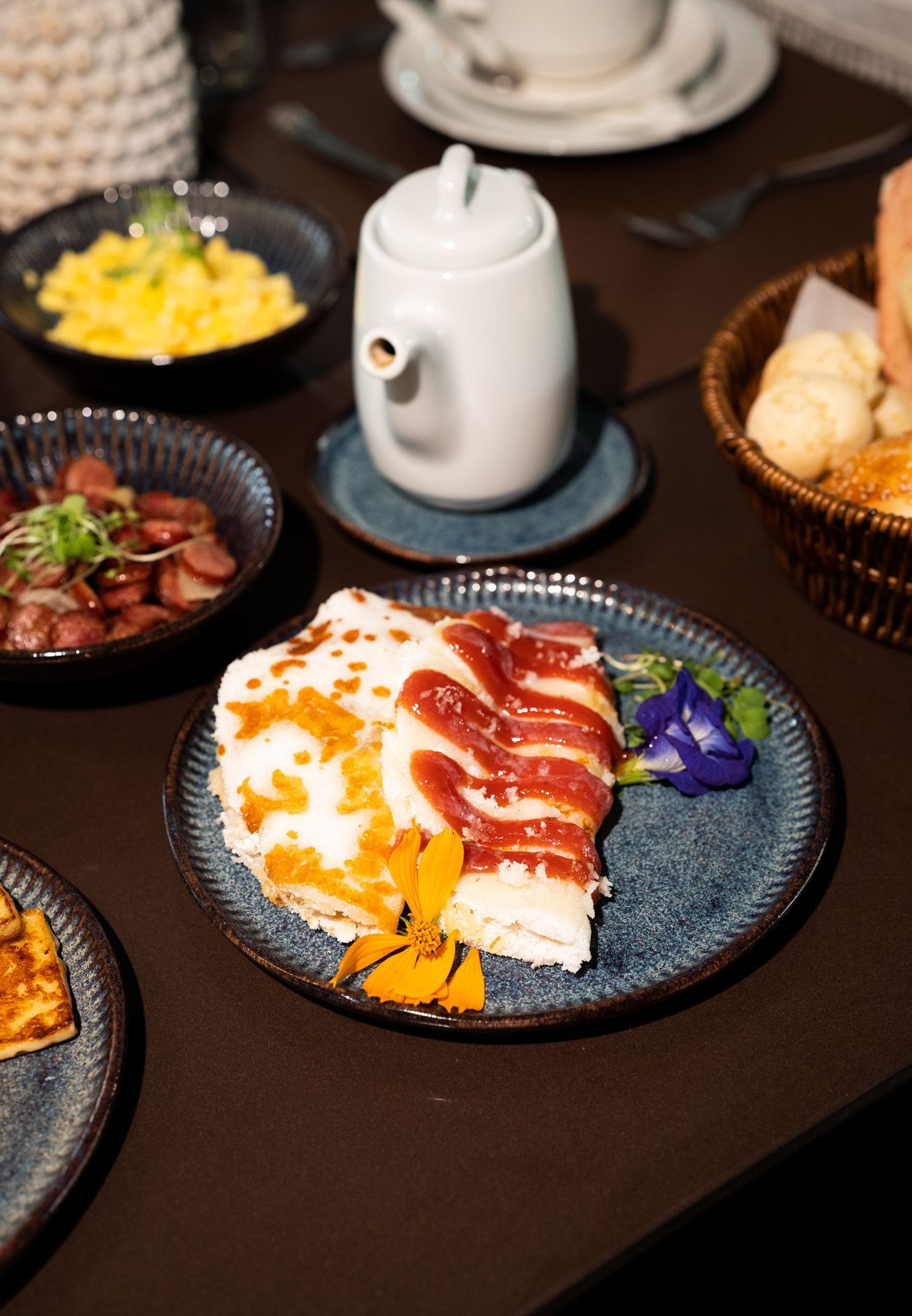 Café da manhã completo: bacon, ovos mexidos, linguiça, pão e bule de chá sobre uma mesa escura.