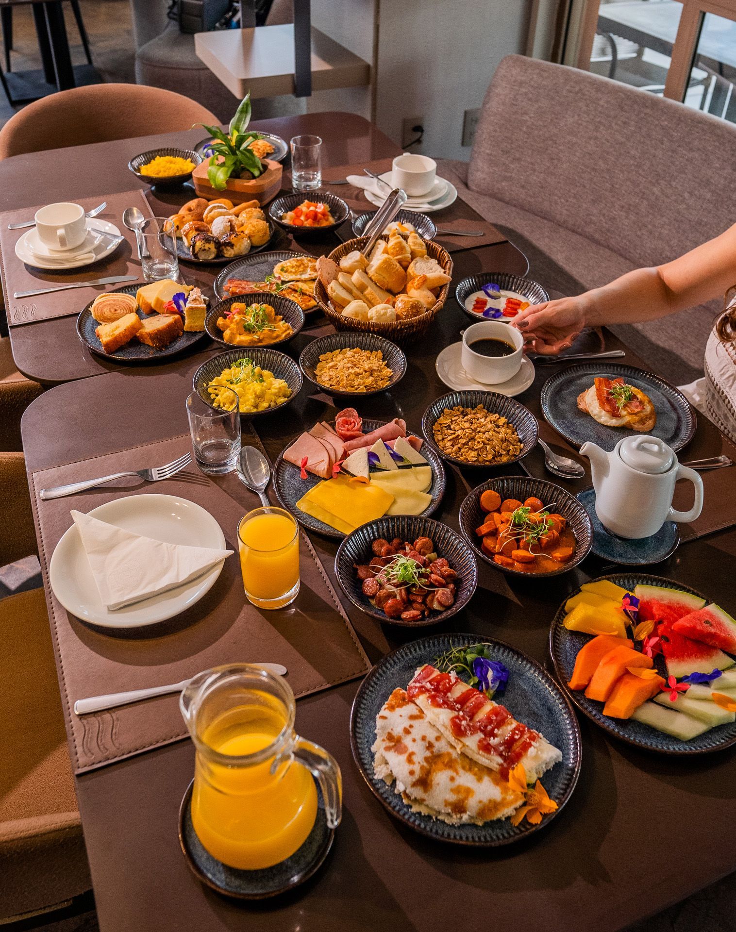 Buffet de café da manhã com diversos pratos: frutas, doces, ovos, carnes e bebidas em uma mesa.