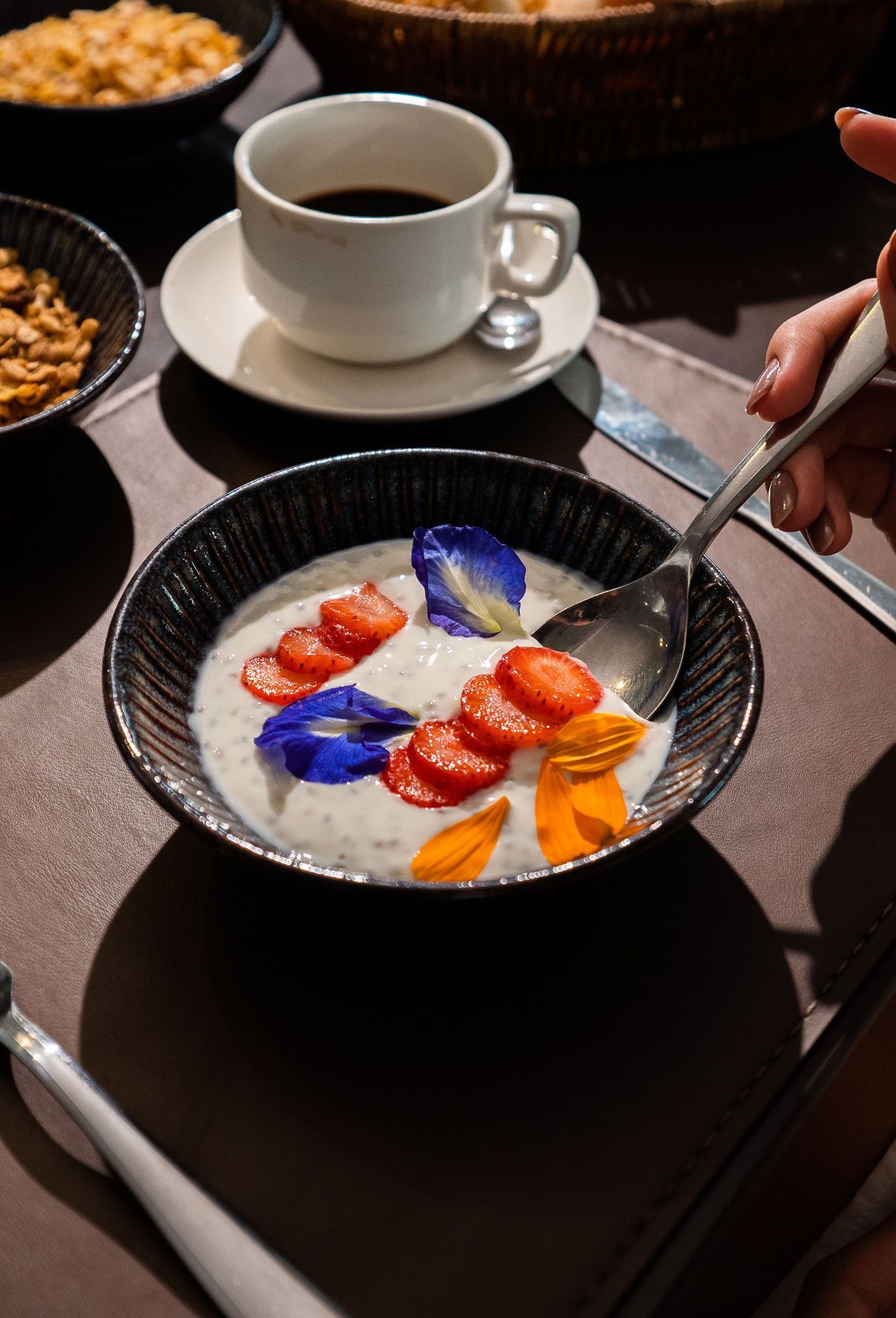 Tigela de pudim com frutas e flores, café e pessoa comendo à mesa.