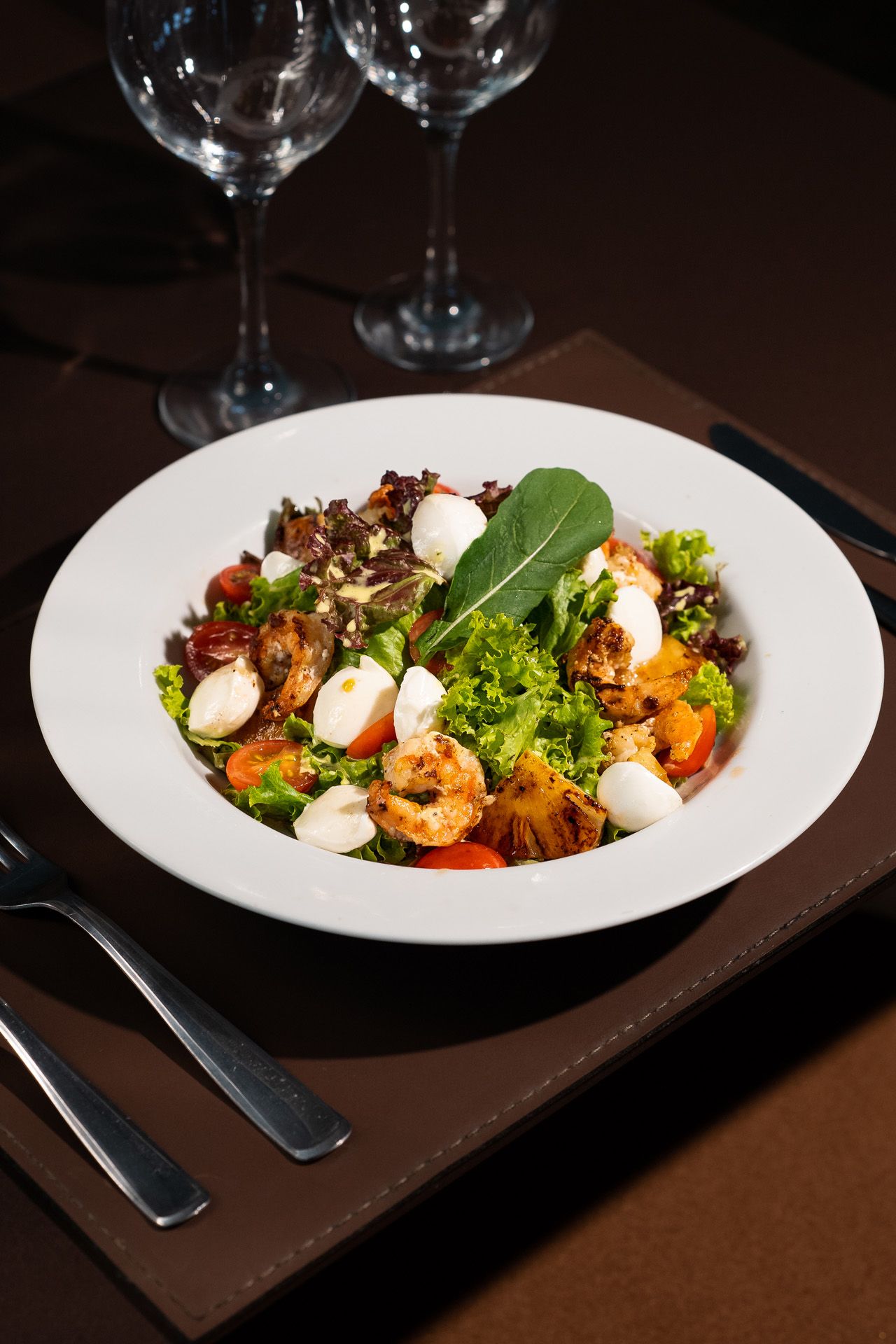 Salada com mussarela, folhas verdes e camarão em um prato branco, servida com taças de vinho e talheres.