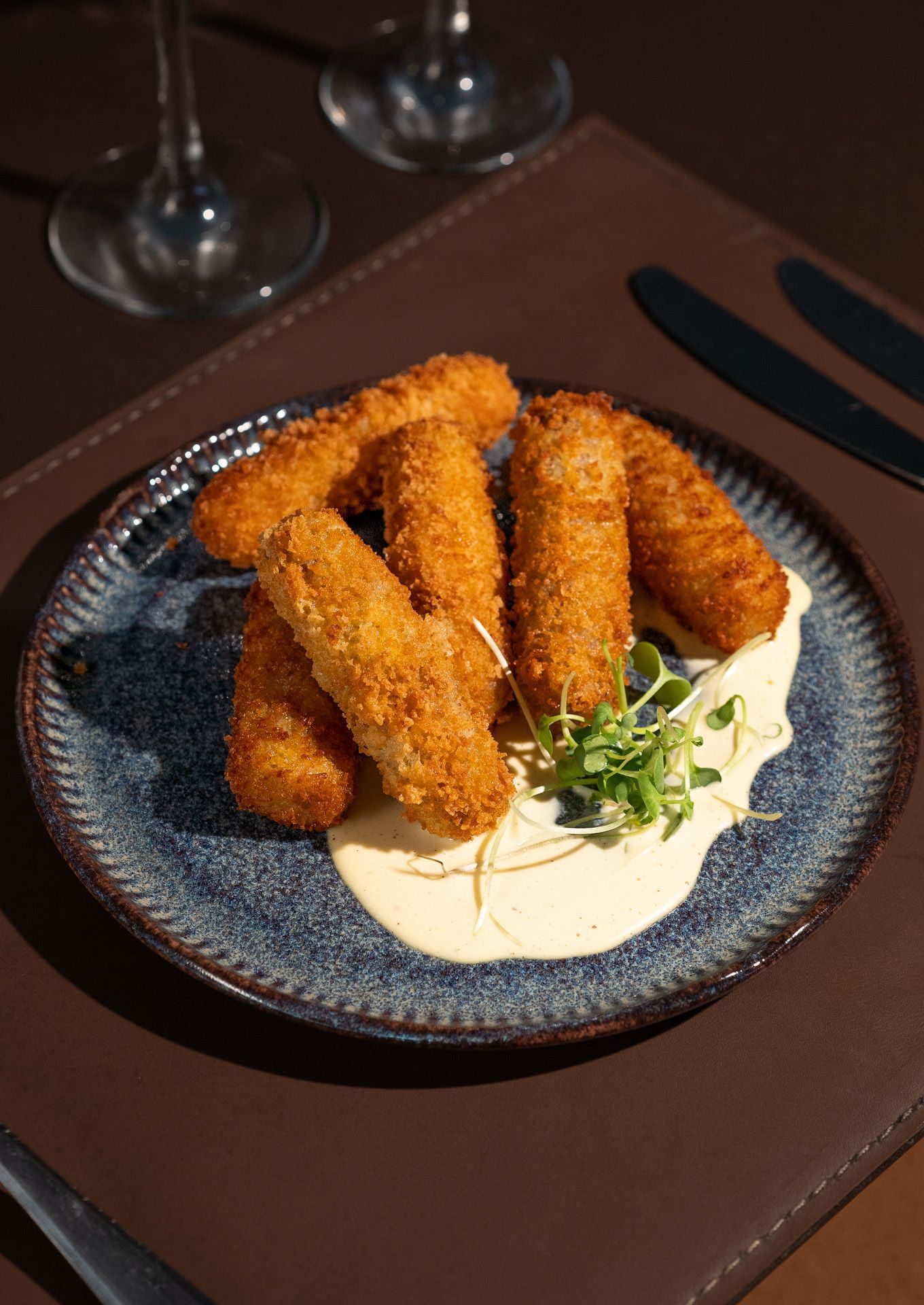 Alimentos cilíndricos, empanados e dourados, servidos em um prato azul com molho cremoso e guarnição, sobre uma superfície marrom.
