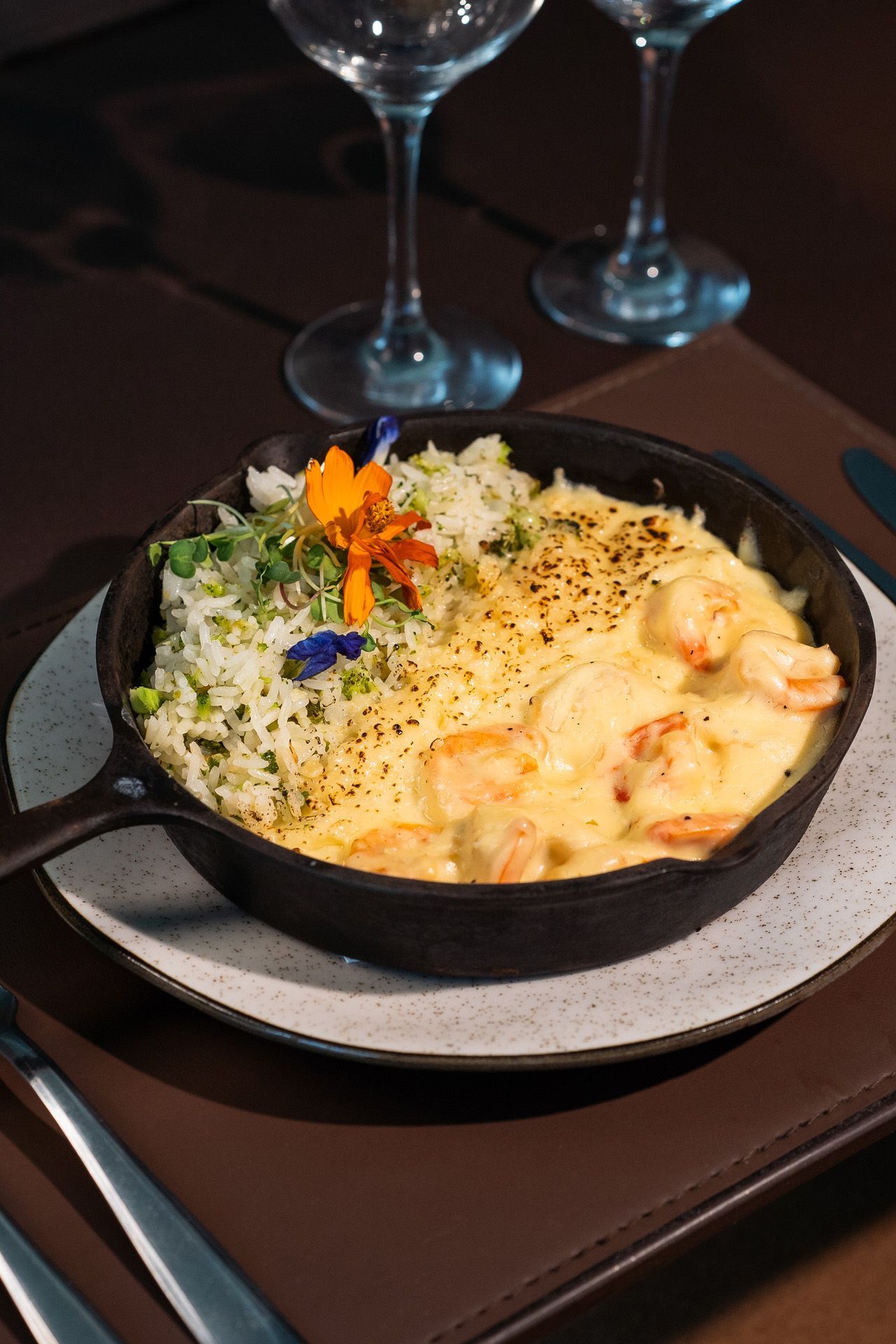 Prato de camarão com arroz servido em uma frigideira de ferro fundido, decorado com flores, em um prato com taças de vinho ao fundo.