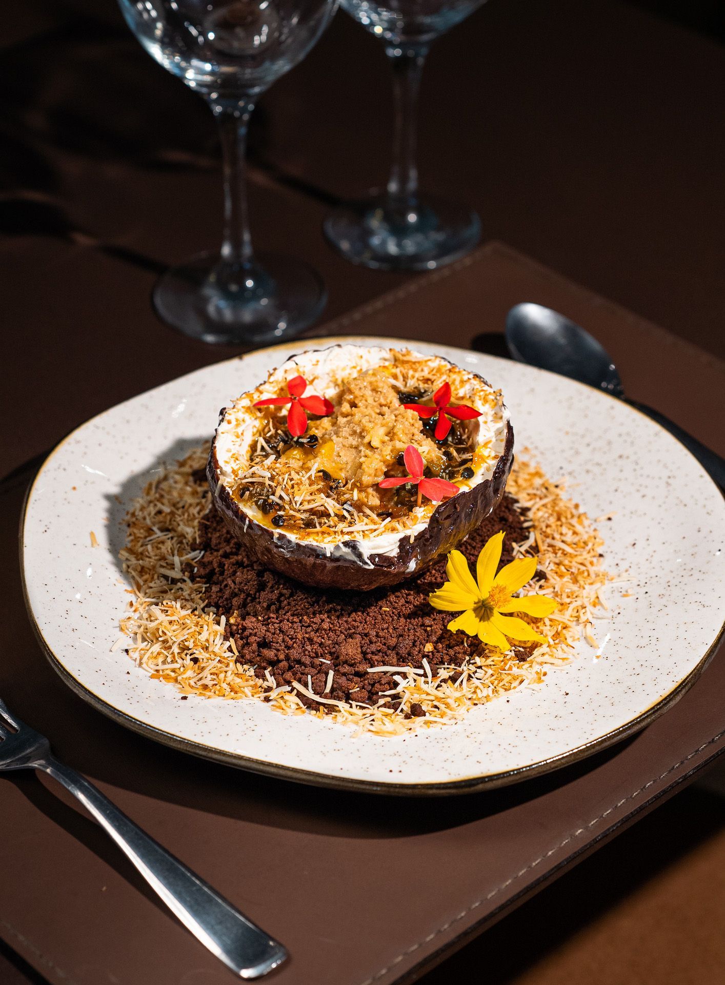 Sobremesa em prato branco: casca de coco recheada com creme e coberturas, rodeada por migalhas de chocolate e coco ralado.