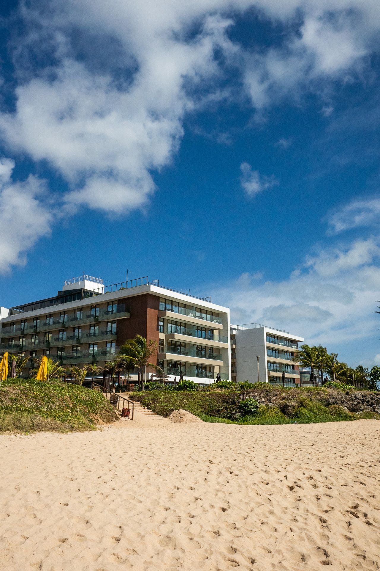 Hotel à beira-mar sob um céu azul com nuvens brancas. Praia de areia em primeiro plano, vegetação ao longo da costa.
