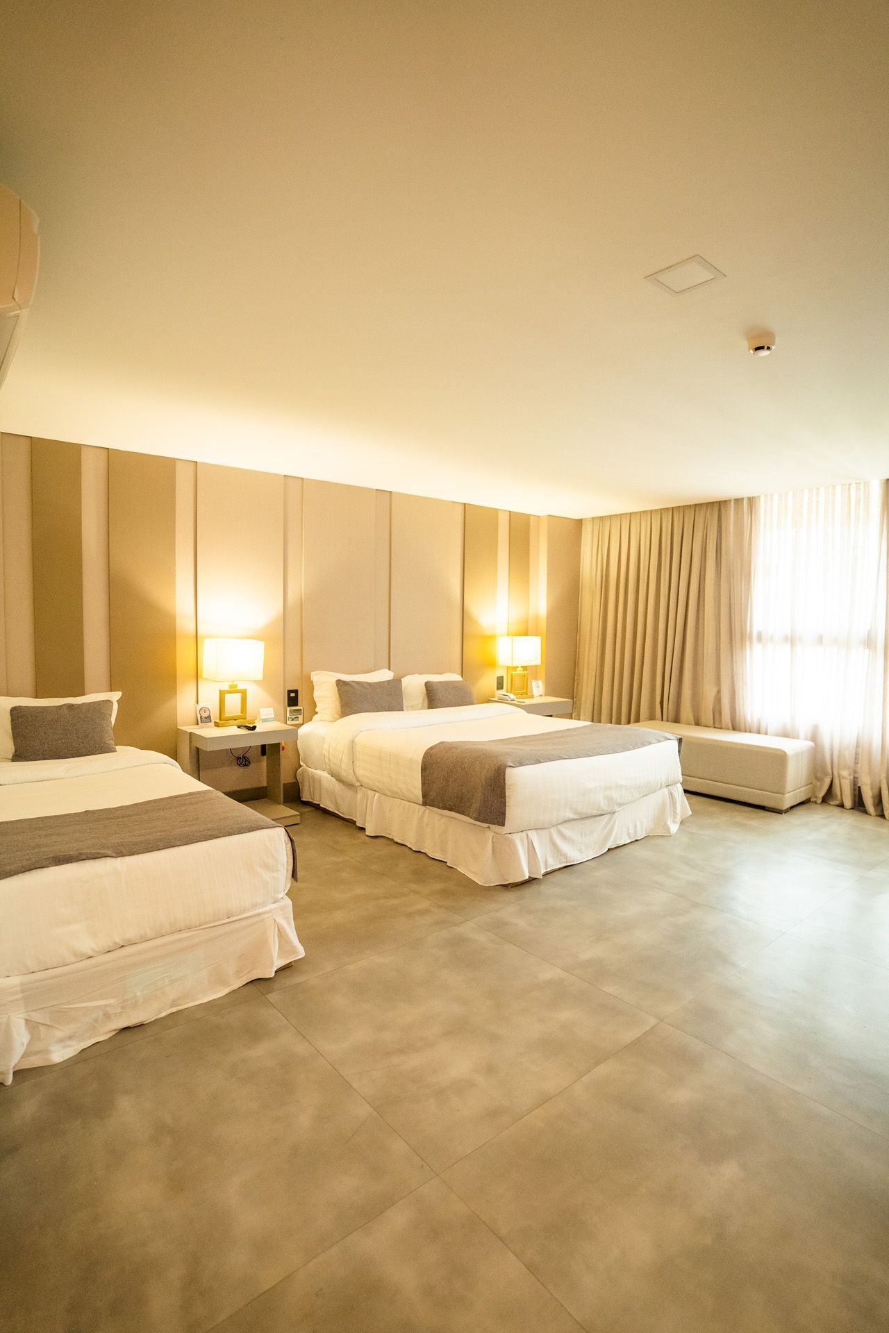 Quarto de hotel com três camas, tons neutros, candeeiros de cabeceira e uma janela com cortinas translúcidas.