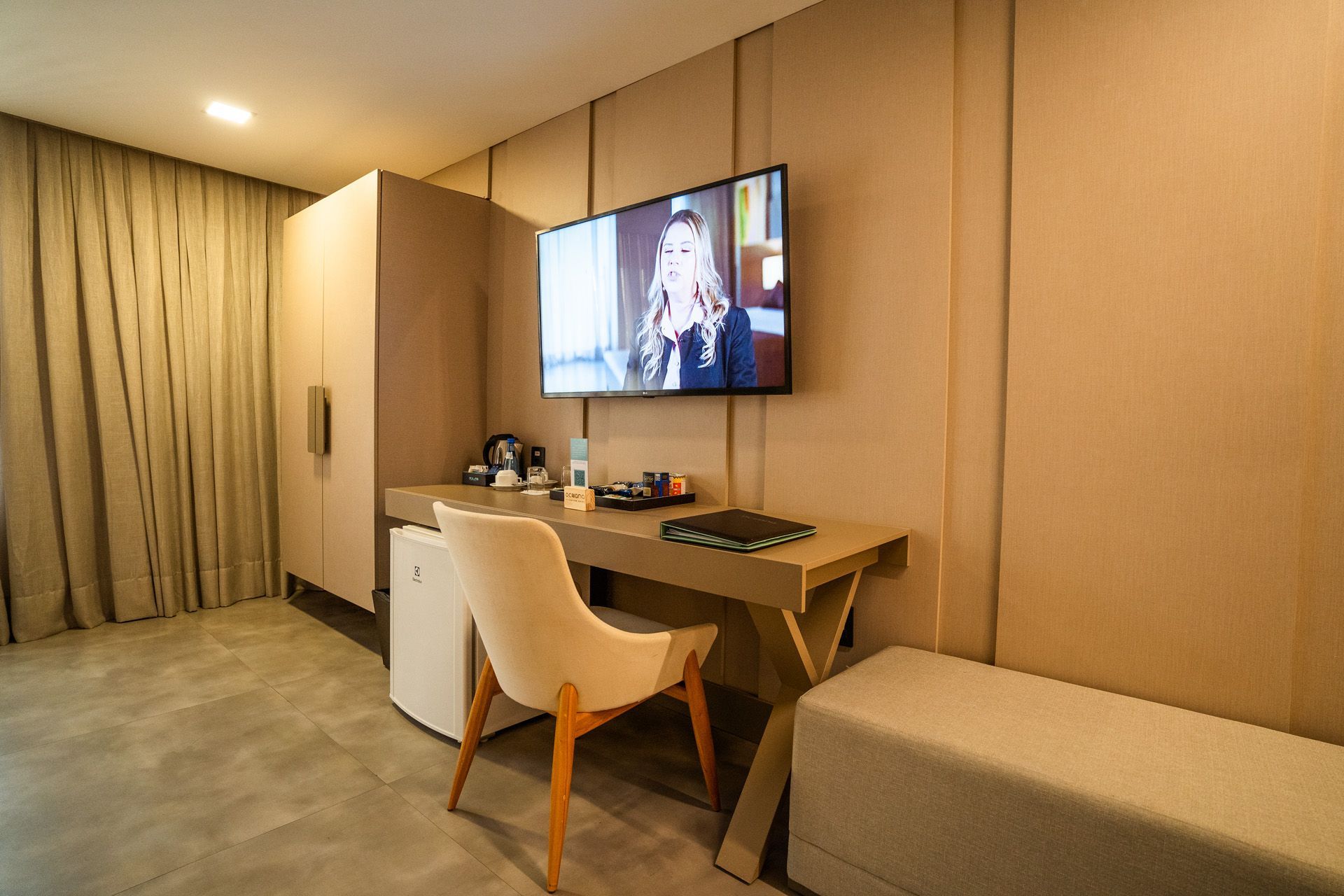Quarto de hotel com escrivaninha, cadeira, TV, armário e banco. Decoração em tons de bege e cinza.