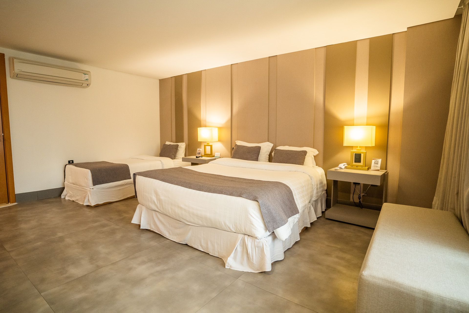 Quarto de hotel com duas camas, tons neutros, mesas de cabeceira, luminárias e ar condicionado.
