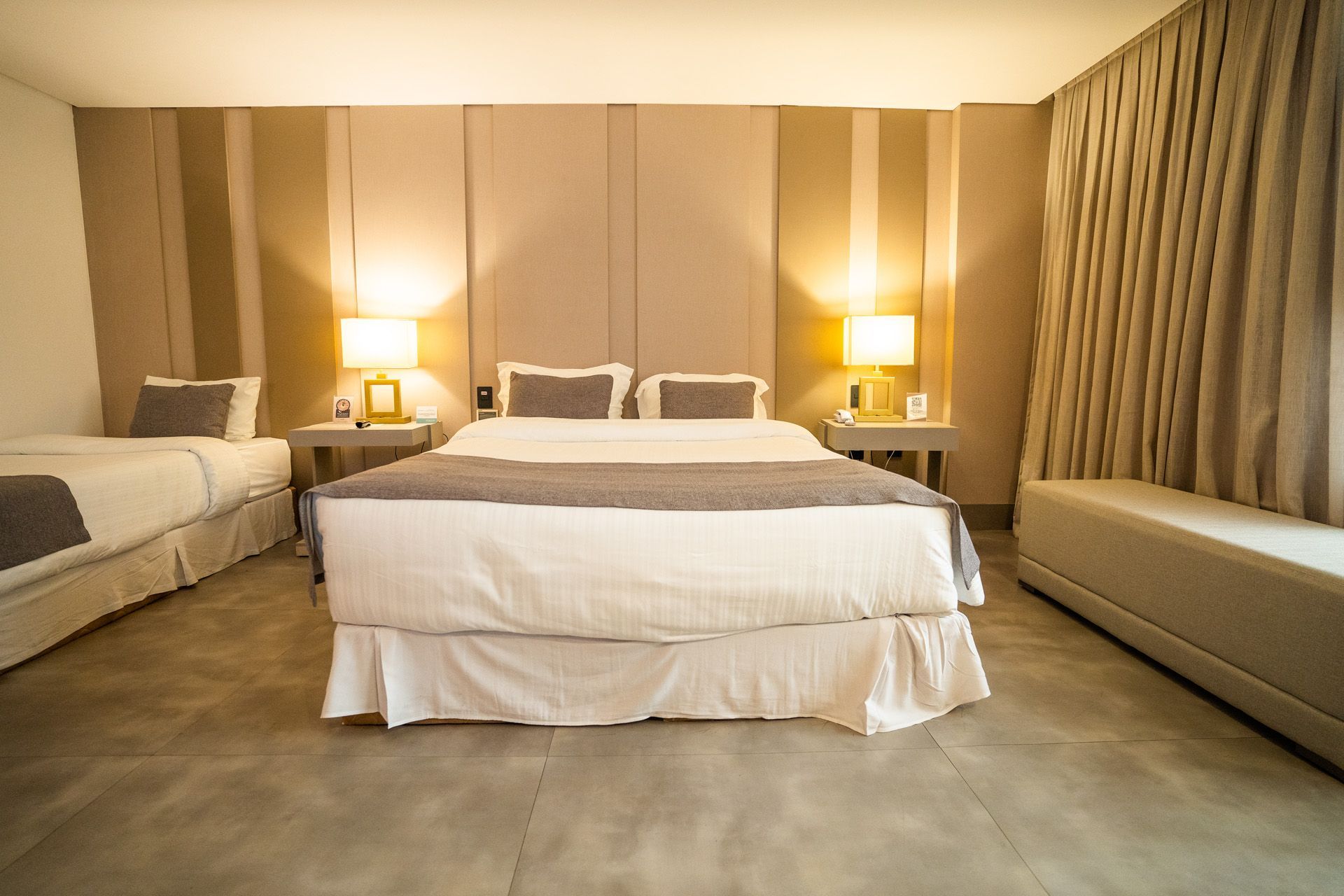 Quarto com duas camas, tons neutros, abajures, cabeceira grande, banco e cortinas.