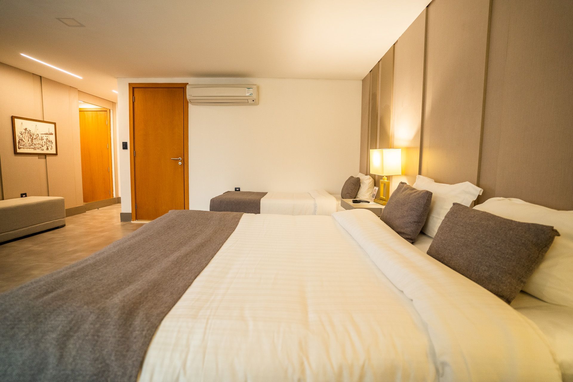 Quarto com duas camas, roupa de cama cinza e branca. Cabeceira grande e porta para o corredor.