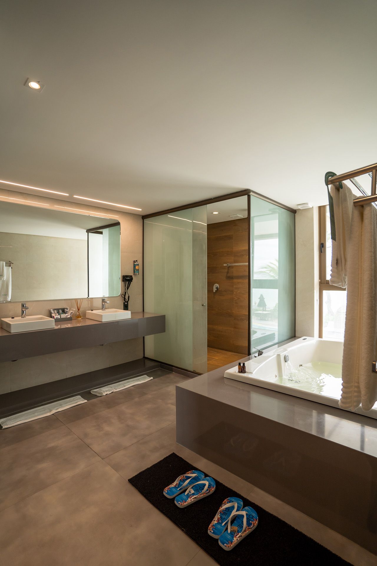 Banheiro moderno com banheira, chuveiro, pia dupla e chinelos no chão.