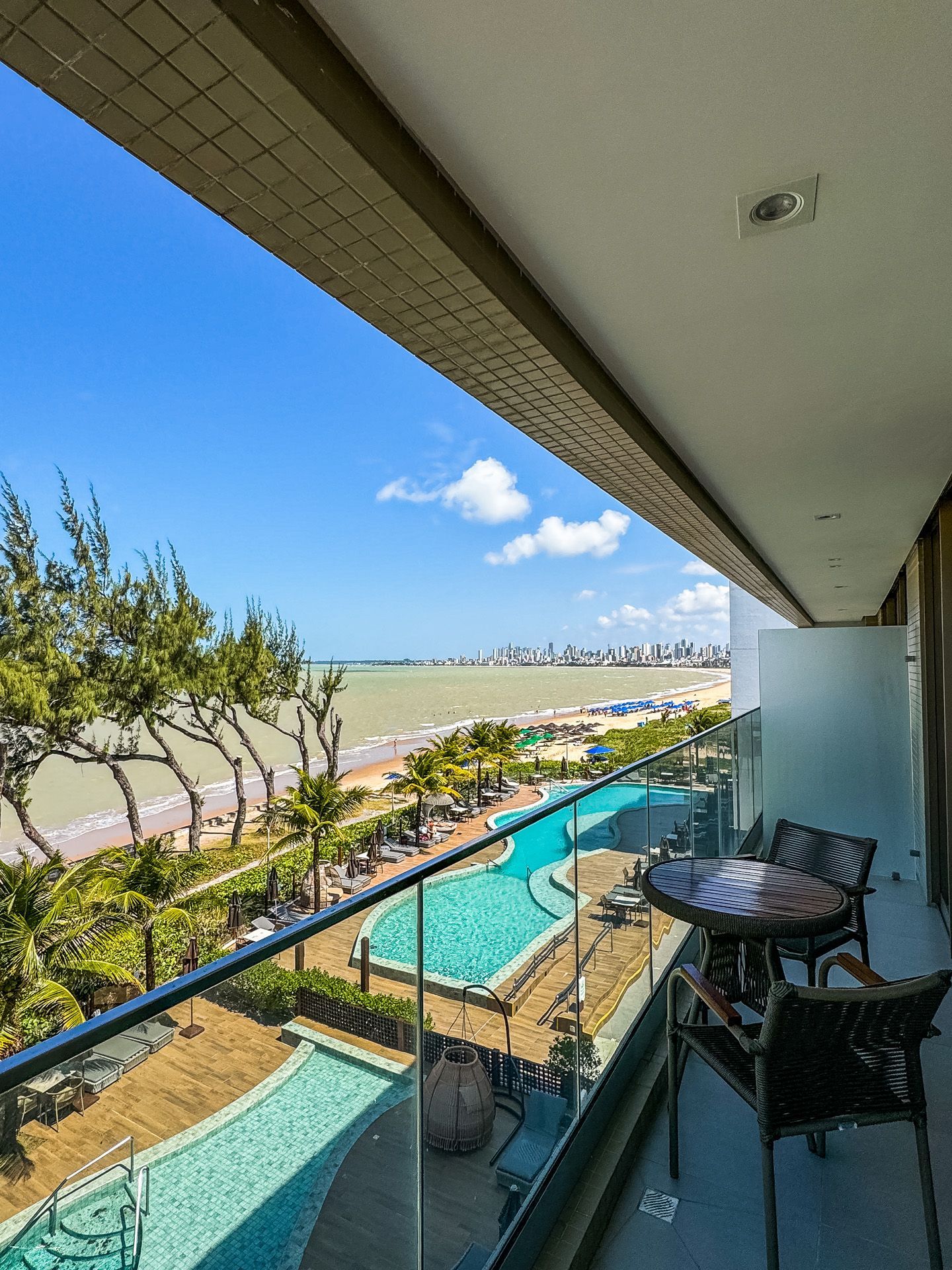 Vista da varanda para a praia, piscina e cidade sob um céu azul. Área de estar externa visível.