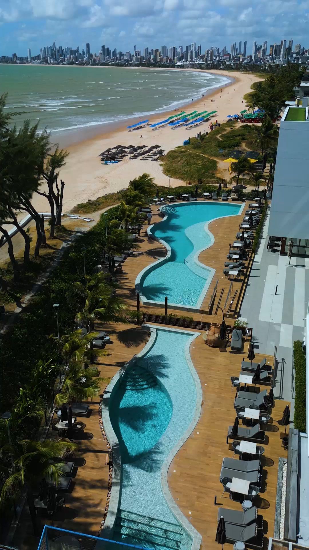 Hotel à beira-mar com piscinas, praia com guarda-sóis e paisagem urbana ao fundo.