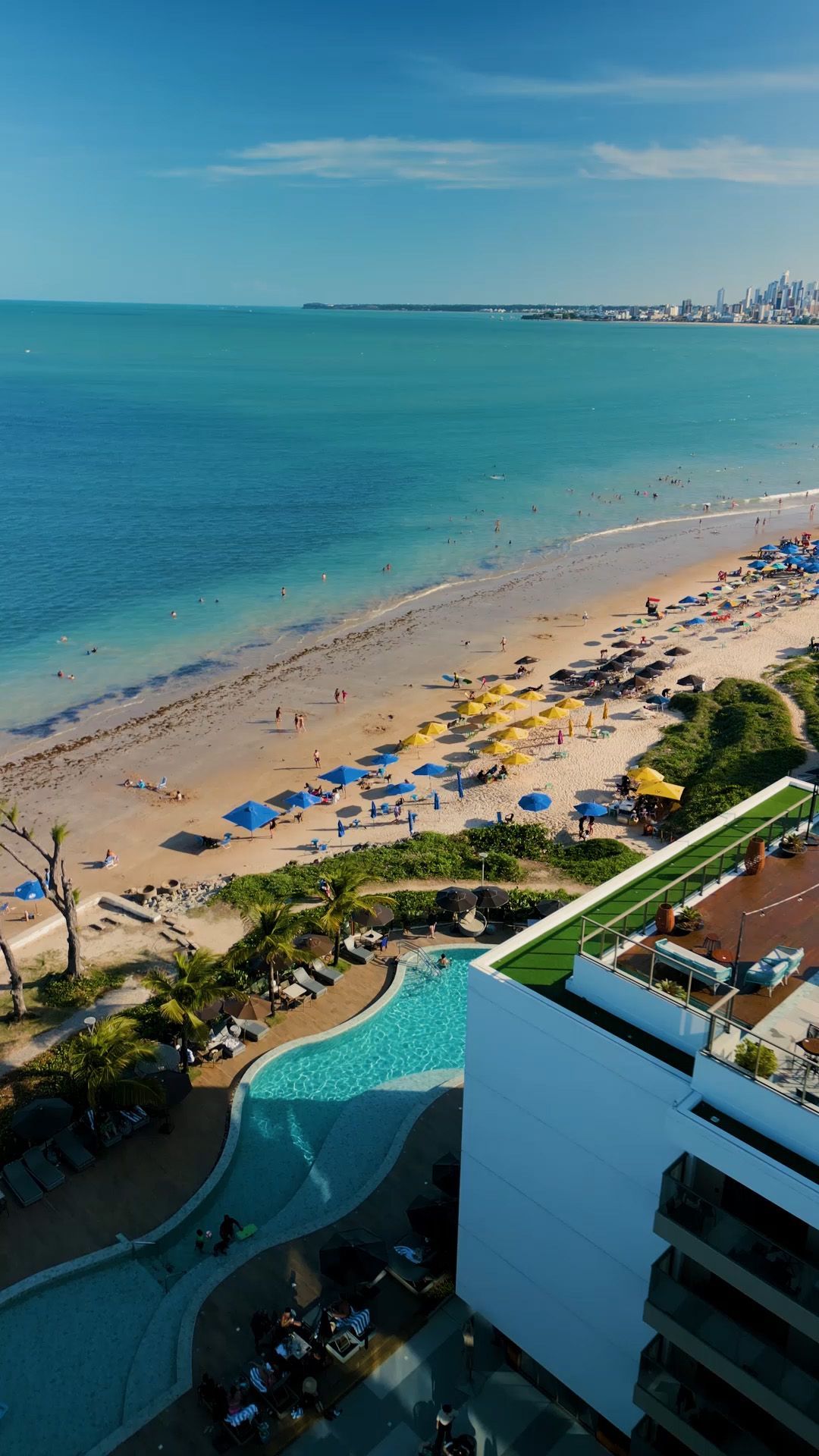 Vista aérea da praia com oceano azul, faixa de areia, guarda-sóis amarelos e azuis e uma piscina turquesa.