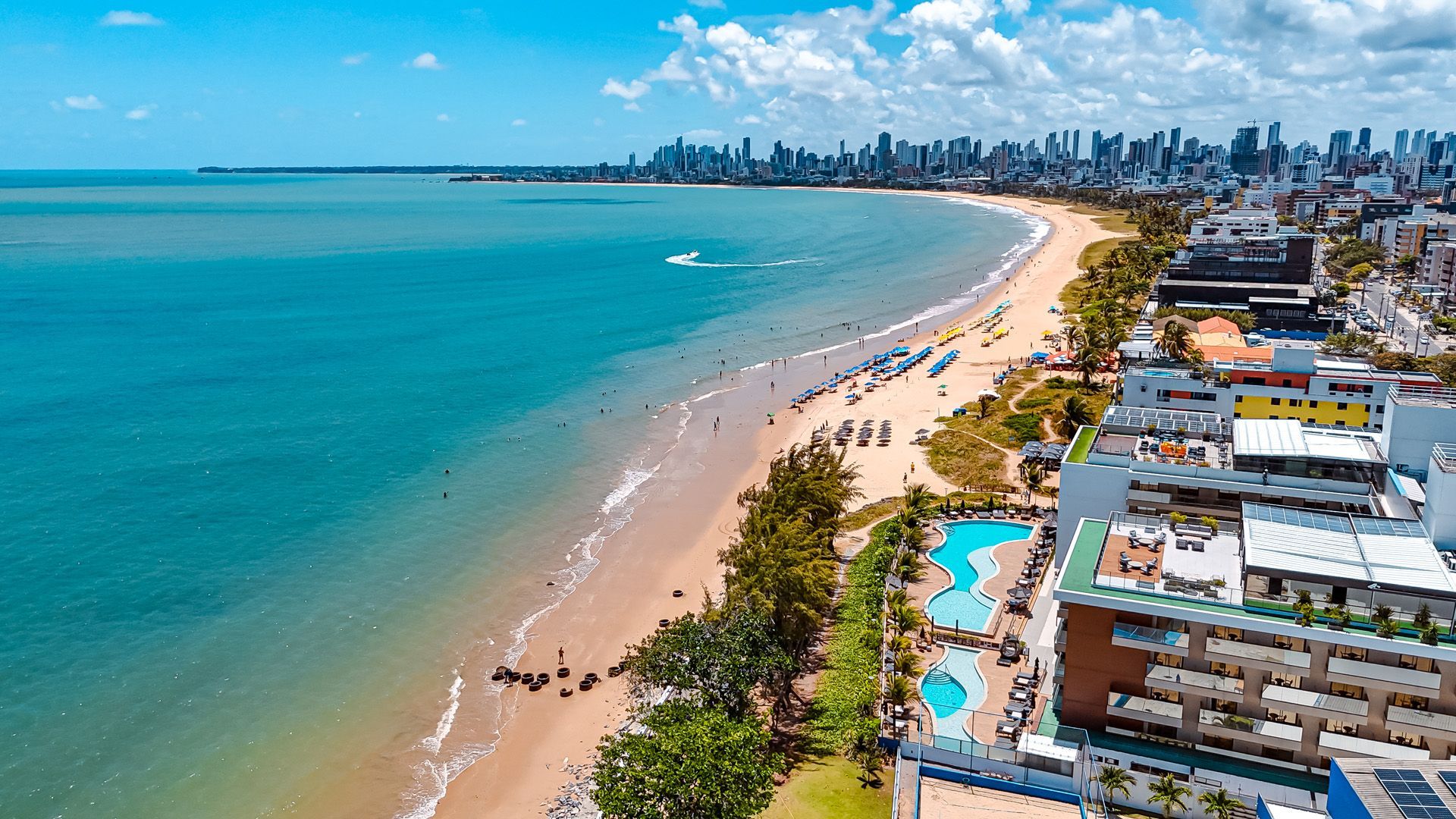 Caiaque azul e remos em uma praia de areia, com cadeiras de praia sob guarda-sóis e o oceano ao fundo.