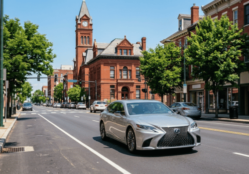 Silver Lexus sedan driving through a historic downtown district with brick buildings, tree-lined streets, and a prominent clock tower in the background.