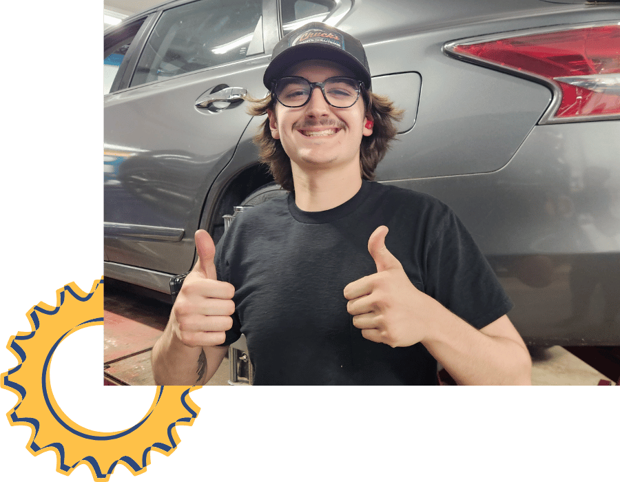 Smiling auto technician giving thumbs up in front of vehicle on lift.