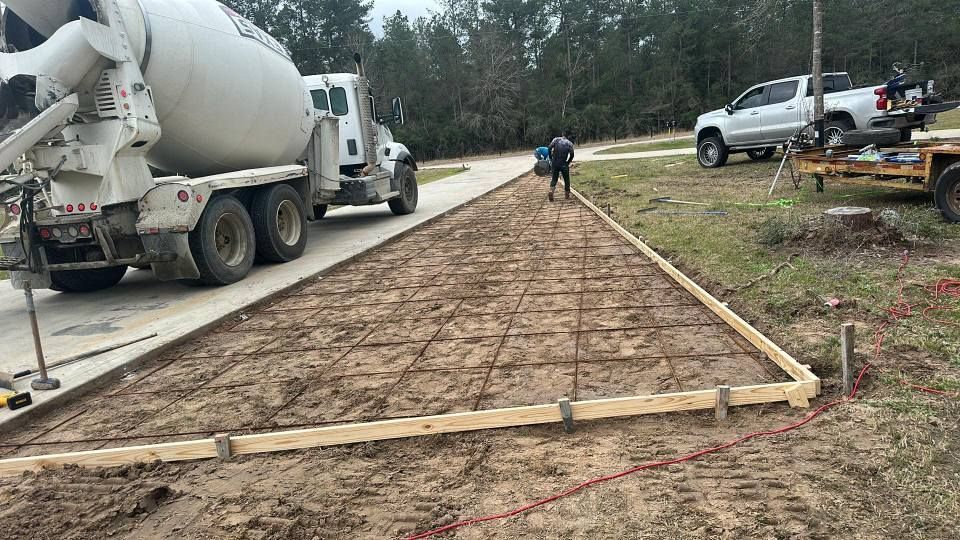 Concrete being poured for a driveway, with cement truck, rebar grid, and workers in a rural setting.
