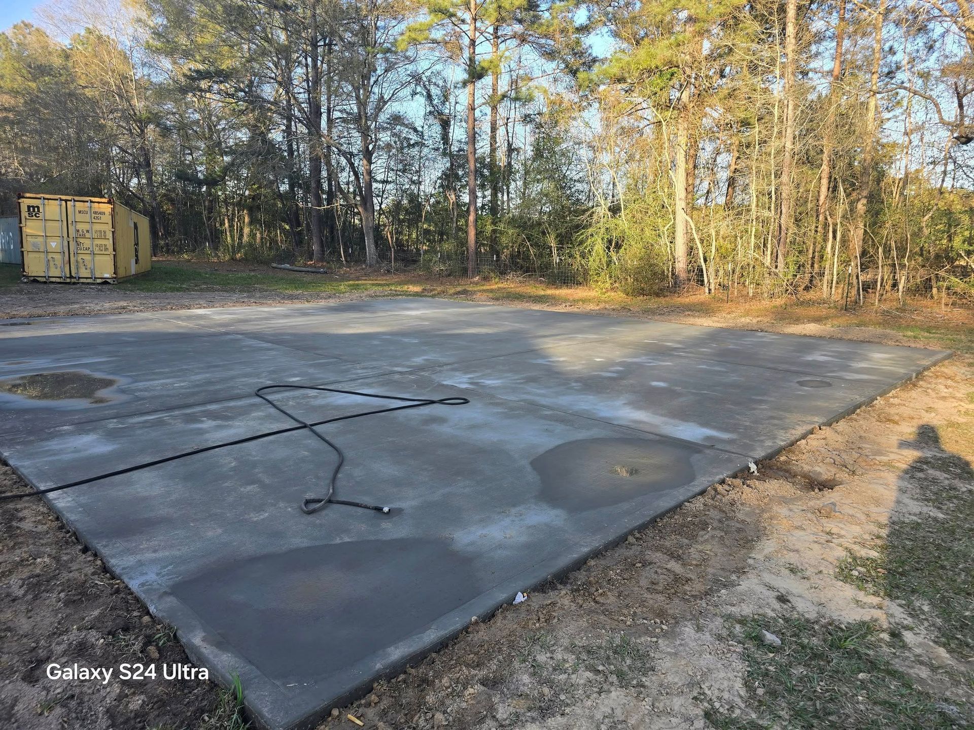 Concrete slab with a hose on it, in a grassy area with trees and a container in the background.