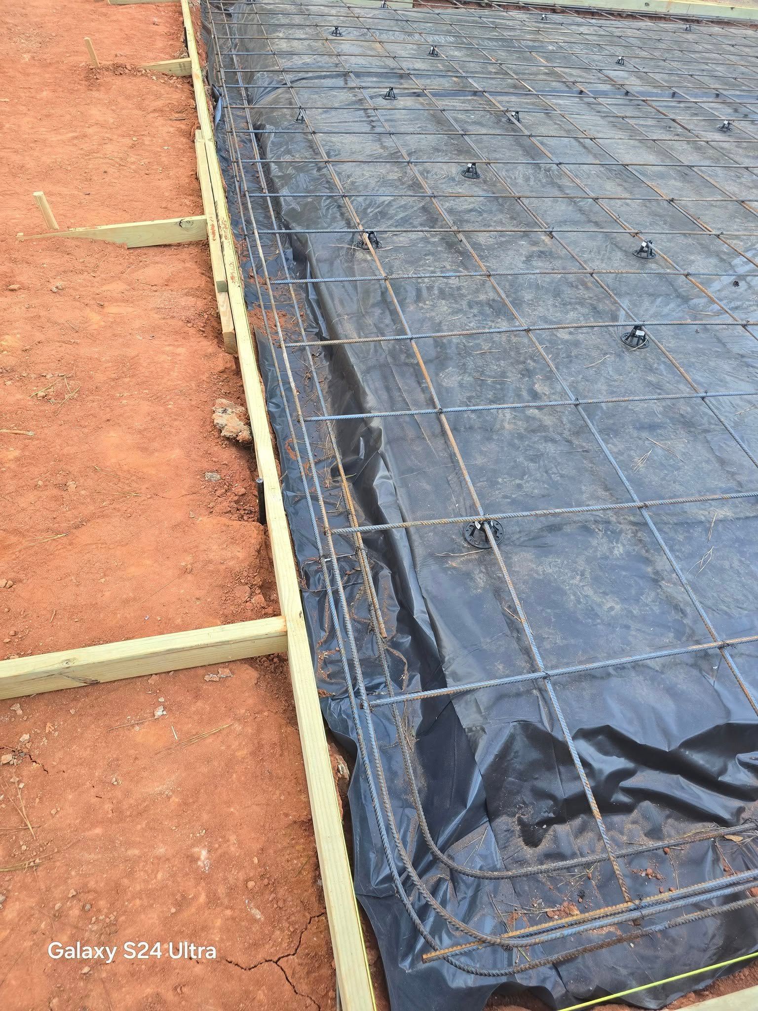 Concrete foundation under construction, with rebar grid over black sheeting within wood forms, set on reddish-brown ground.