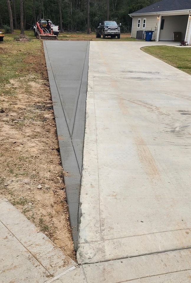 Newly poured concrete driveway extension, gray, adjacent to existing driveway, brown soil on one side.