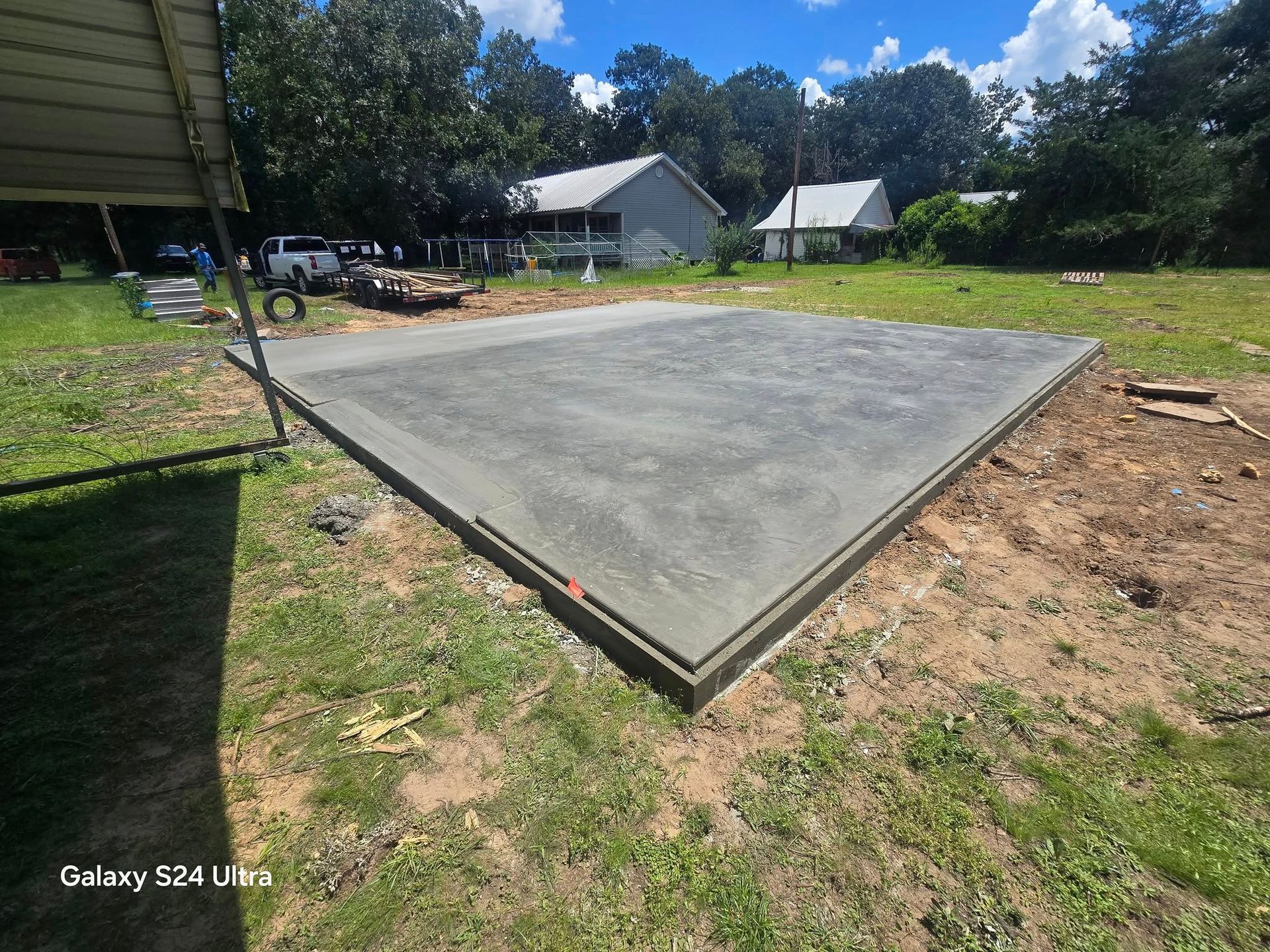 Newly poured concrete foundation in a grassy yard, surrounded by dirt and construction materials, under a blue sky.