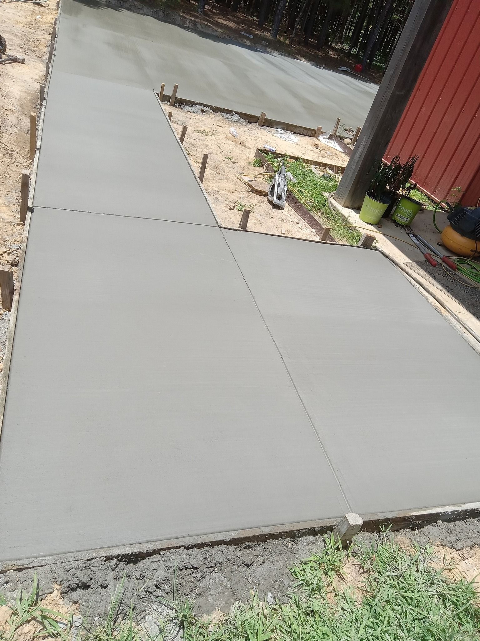 Freshly poured concrete walkway, gray, with wood forms; outdoor setting near a red building.