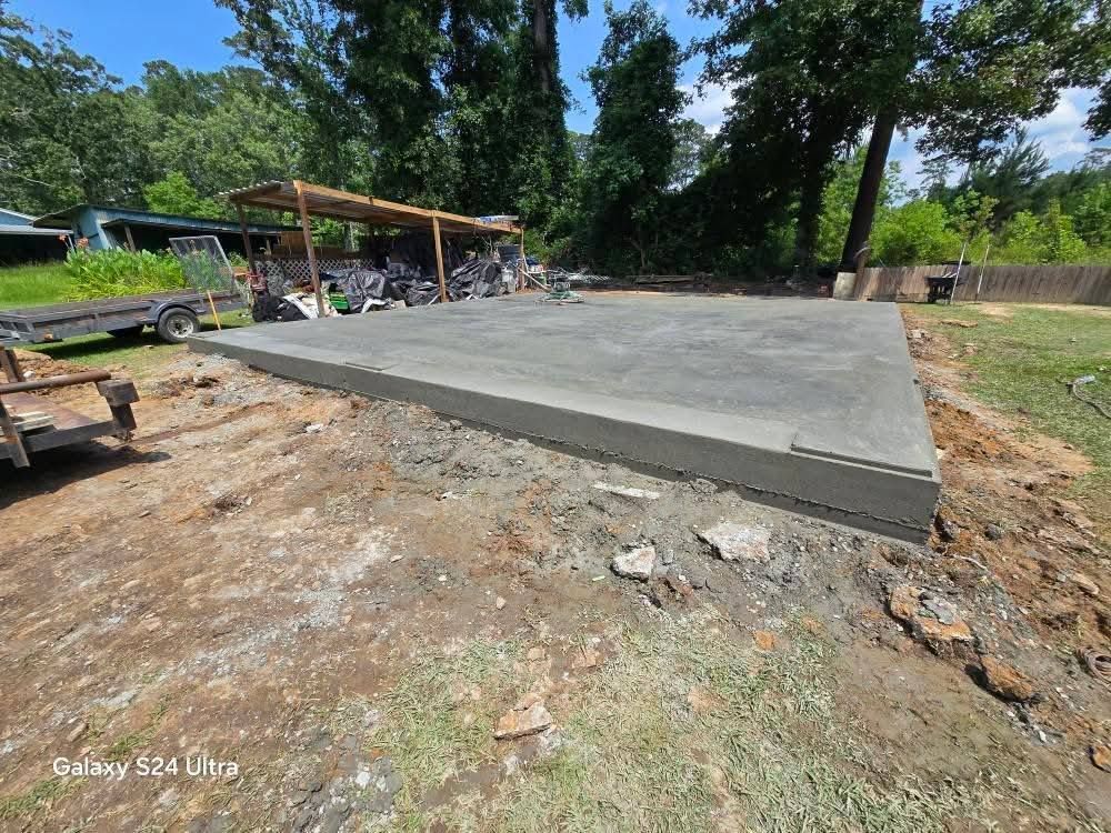 Newly poured concrete slab in an outdoor setting, with a dirt foreground and a structure in the background.