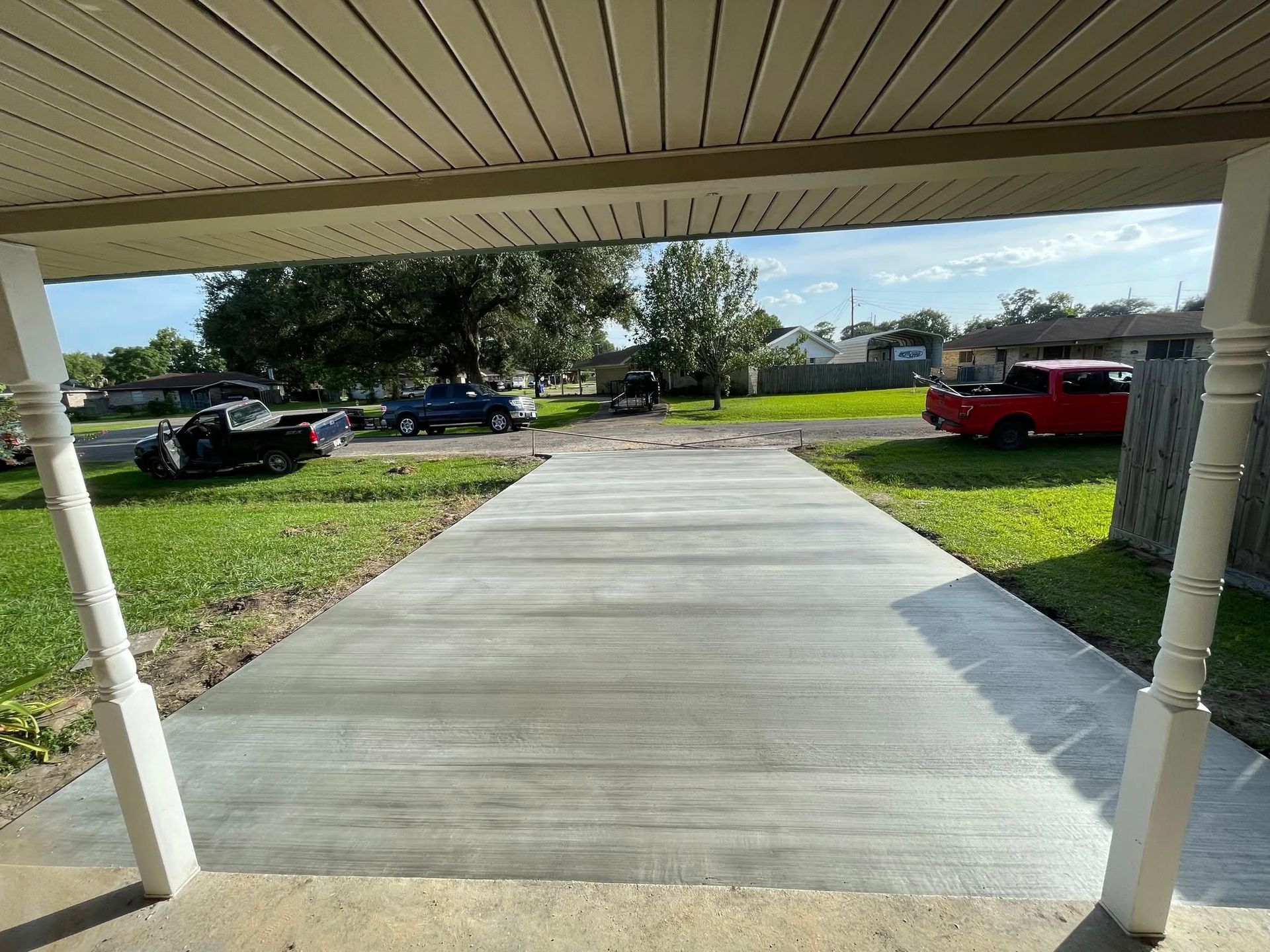 View from a porch of a newly poured concrete driveway, with grass and parked vehicles visible.