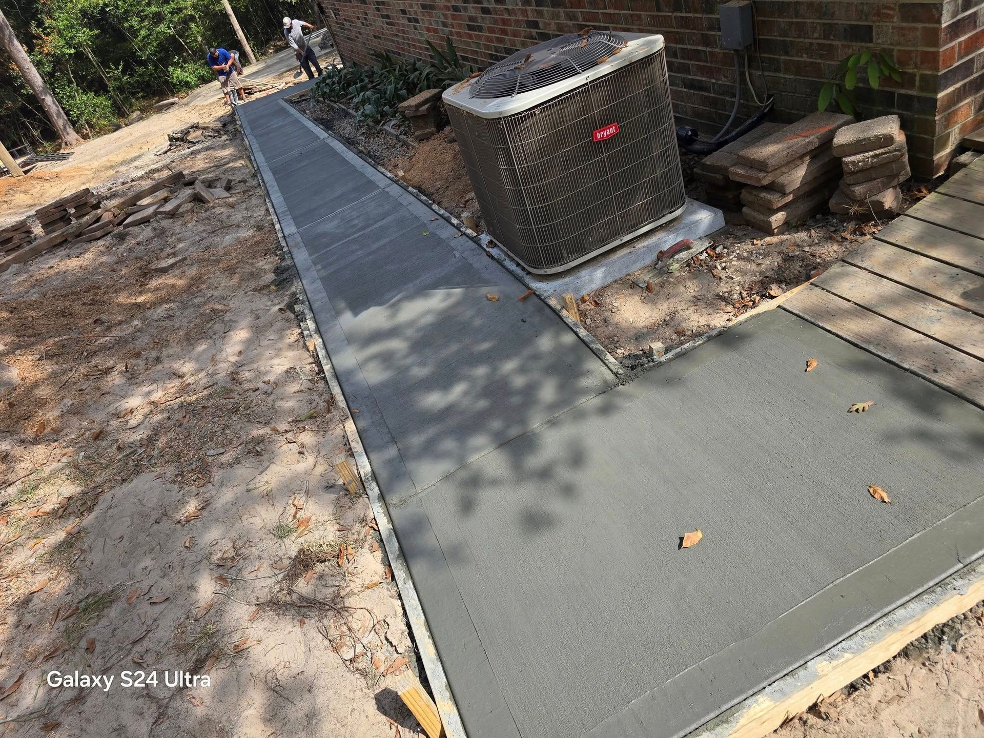 Freshly poured concrete sidewalk next to a building, with an AC unit and bordering stones.