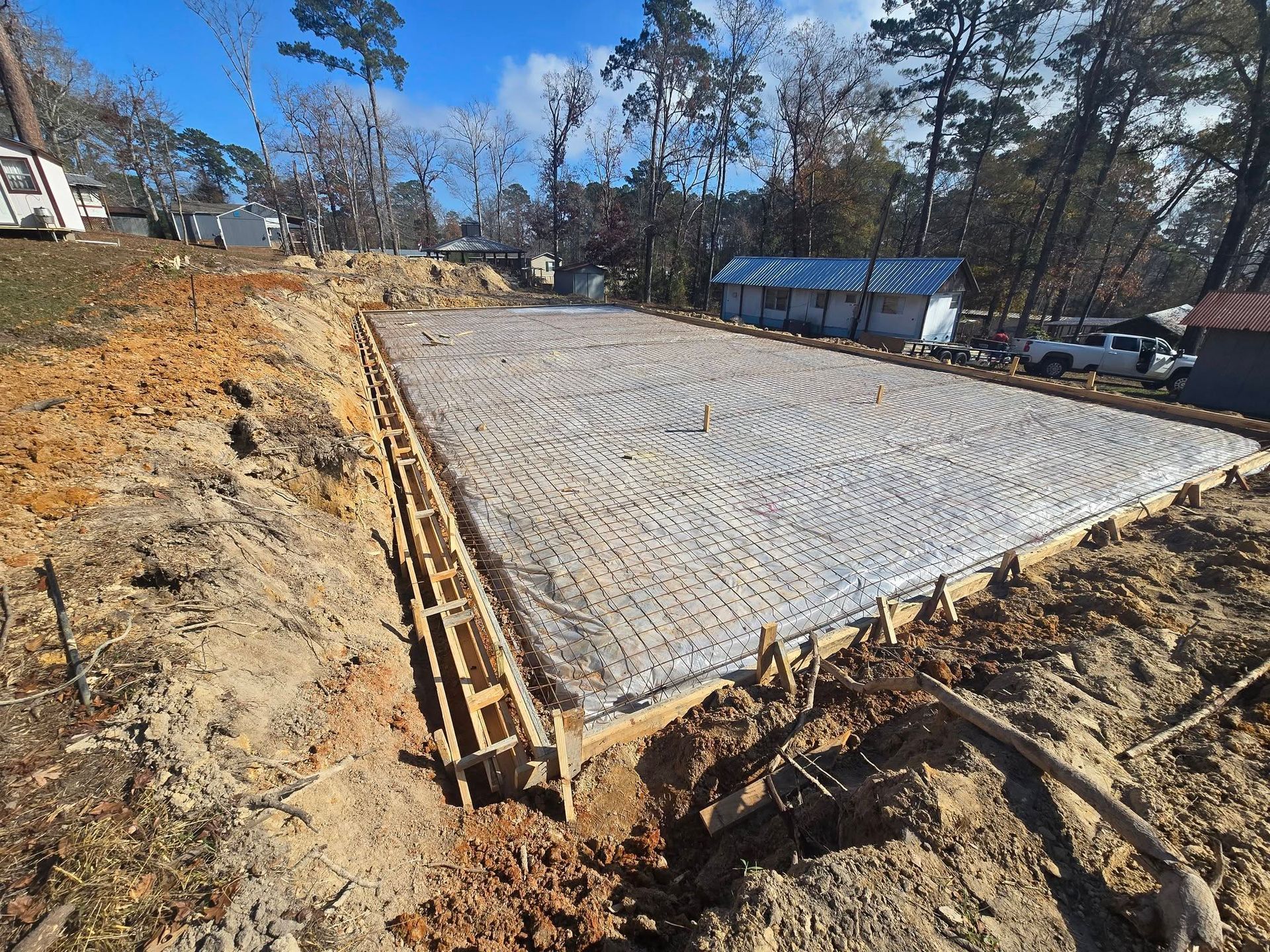 Concrete foundation poured, wooden frame, dirt surrounding, trees in background, sunny day.