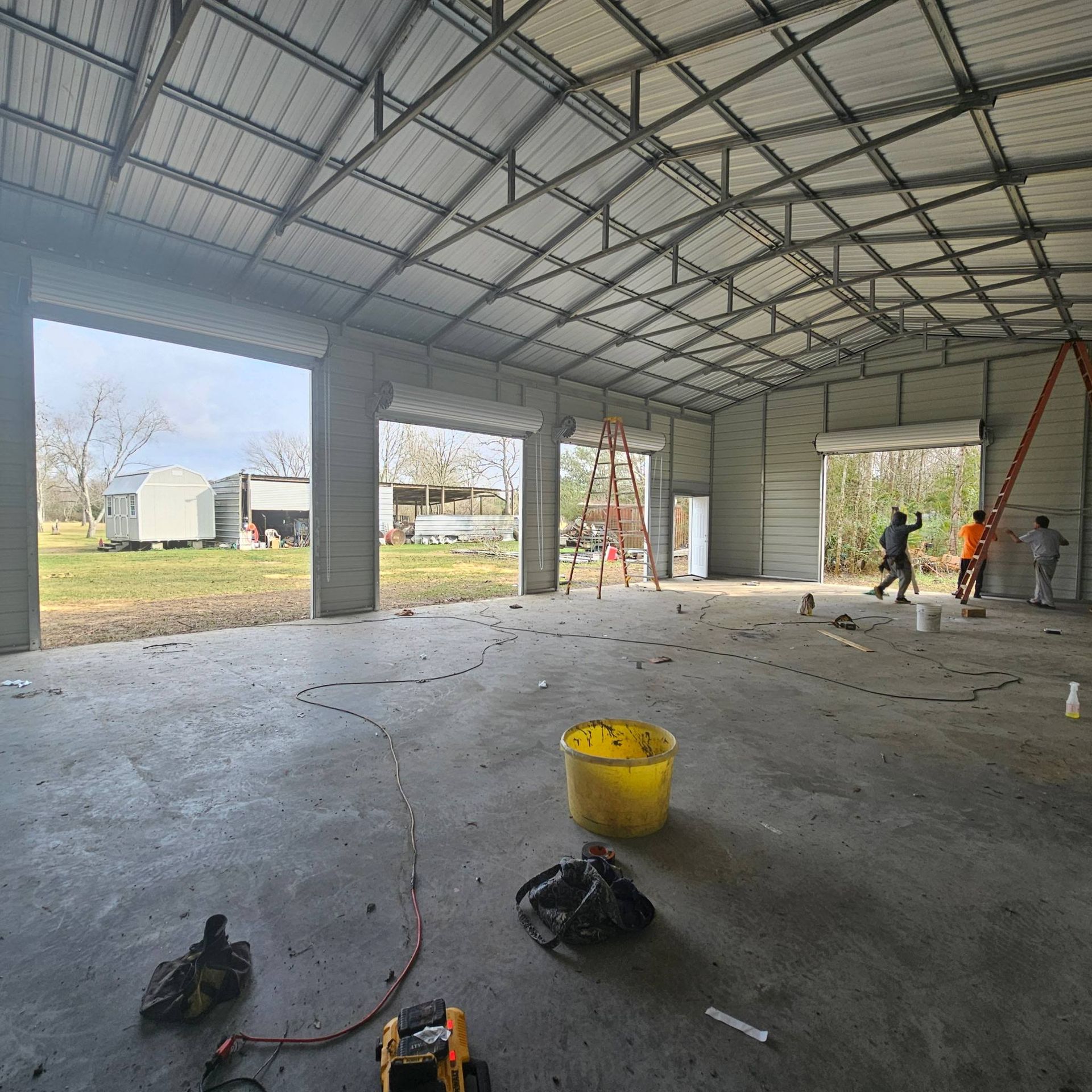 Interior of a metal building with open garage doors. Workers are inside. A yellow bucket and debris are on the floor.