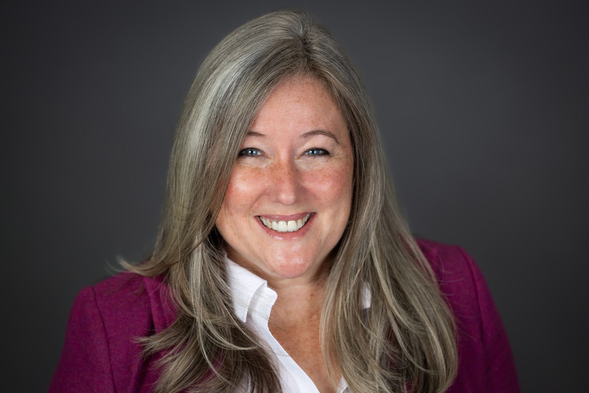 Woman with long gray hair smiling, wearing a magenta blazer and white shirt, against a gray backdrop.