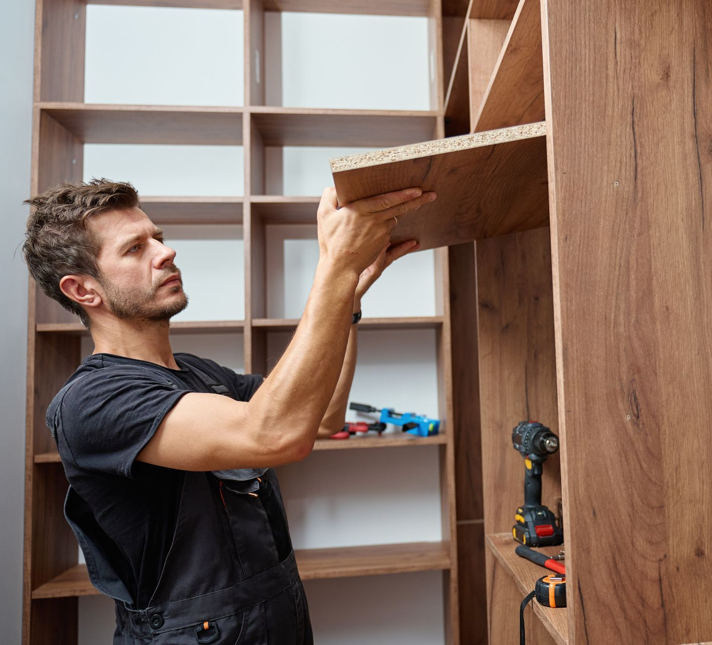 Man installing a shelf in a wooden bookcase, tools visible.