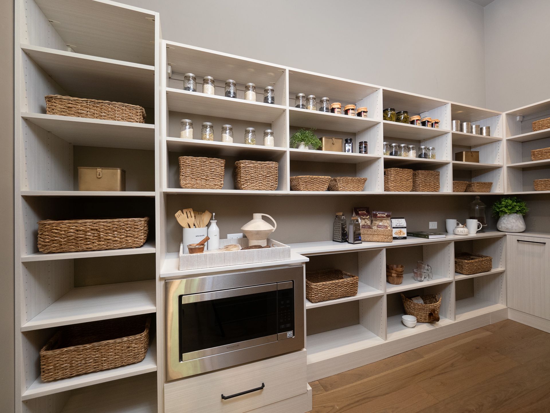 White pantry with open shelving, baskets, and a microwave built-in; wooden floor.