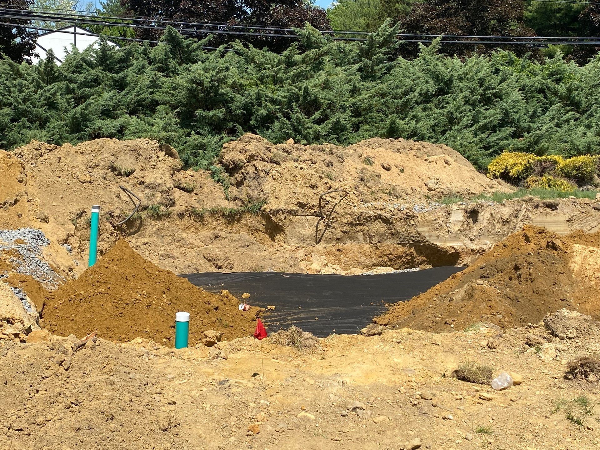 Construction site with a dirt excavation, black liner, and green pipes in a sandy area near shrubs