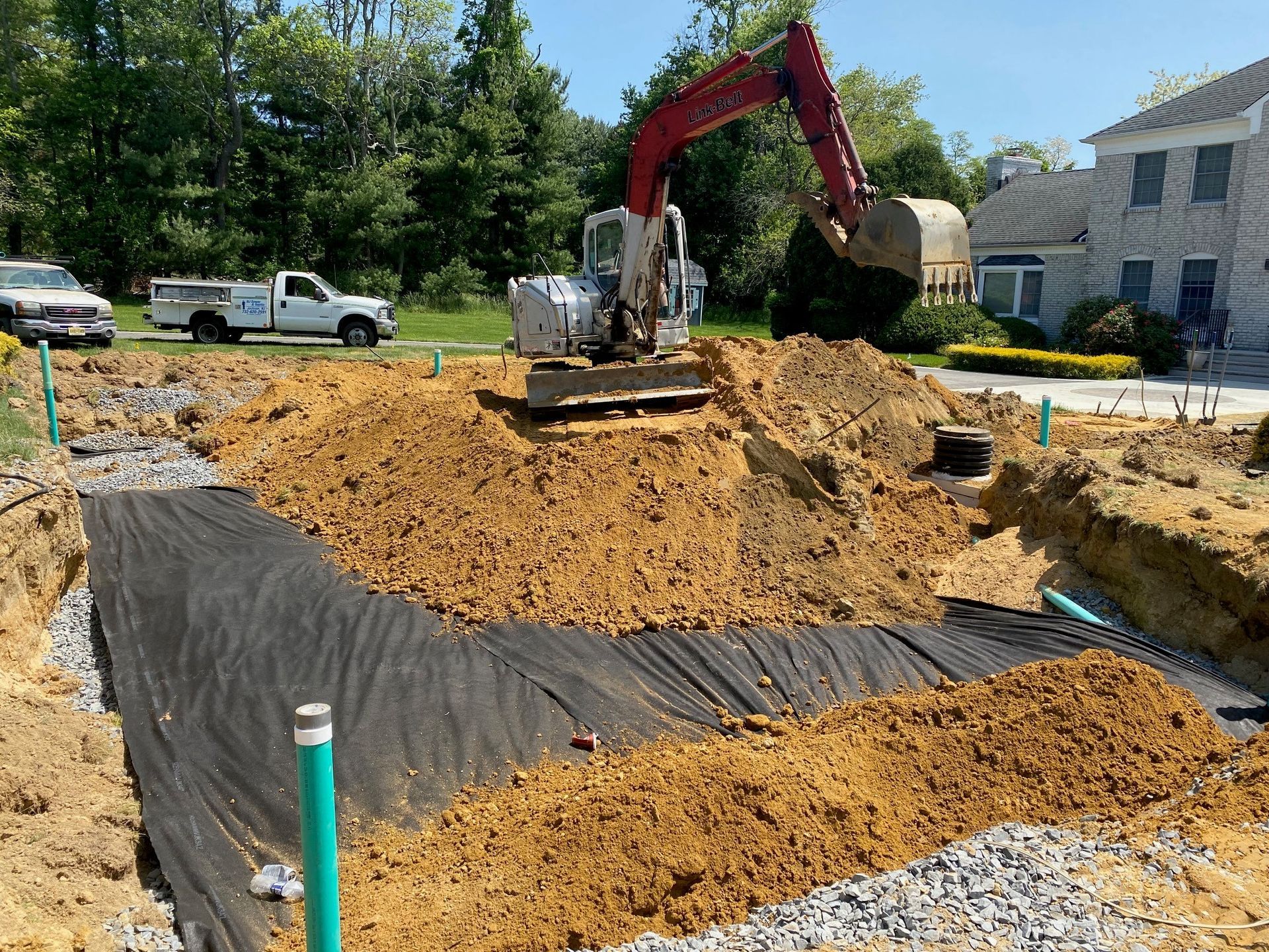 Excavator digging a large hole at a construction site with black tarp lining and dirt piles nearby