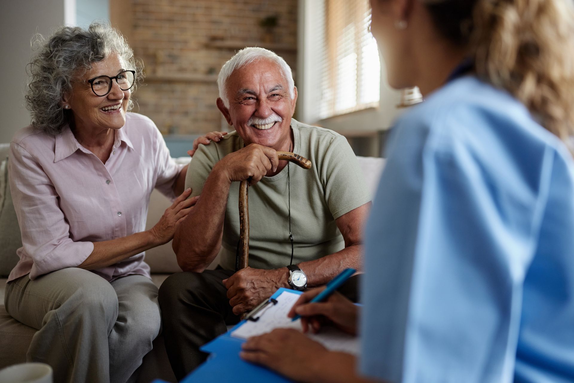 An elderly couple is sitting on a couch talking to a nurse.