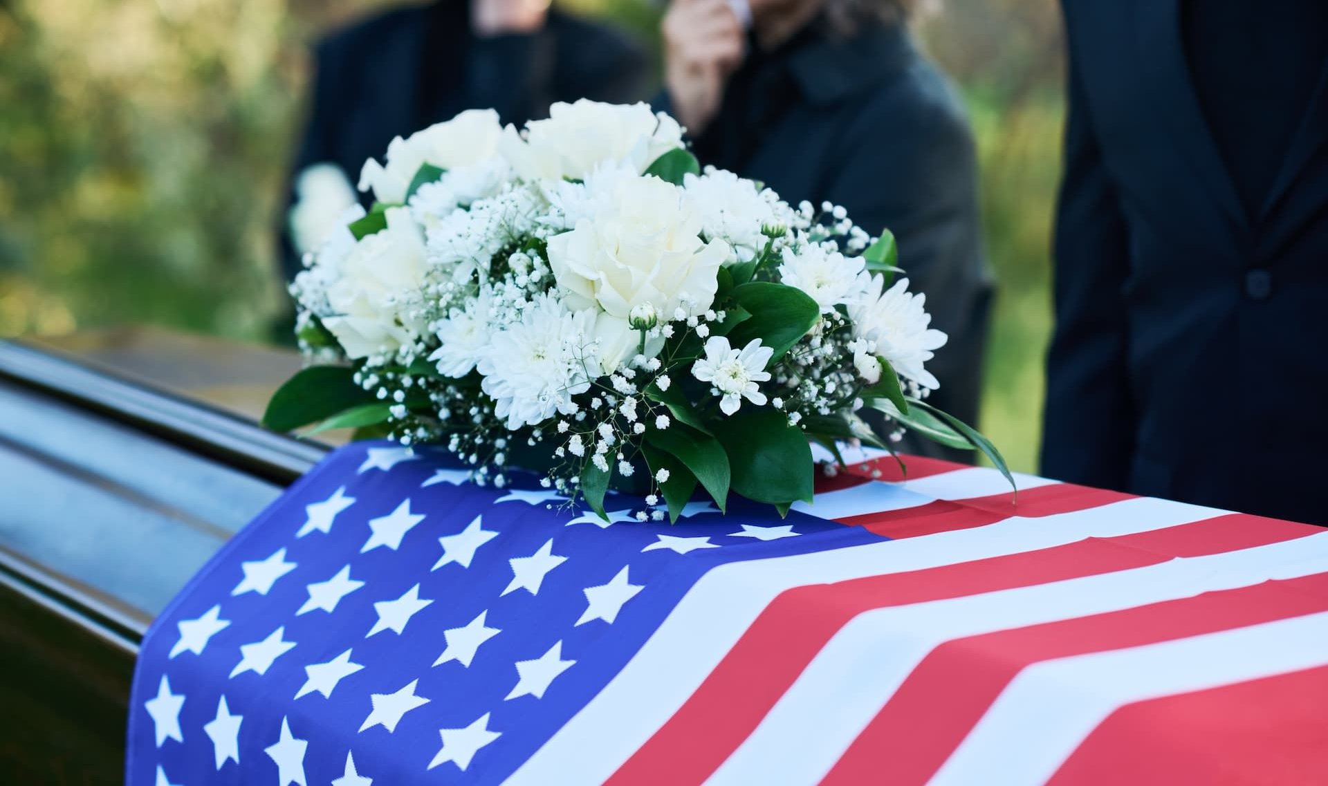 A casket draped with the American flag, topped with white flowers; mourners in the background.