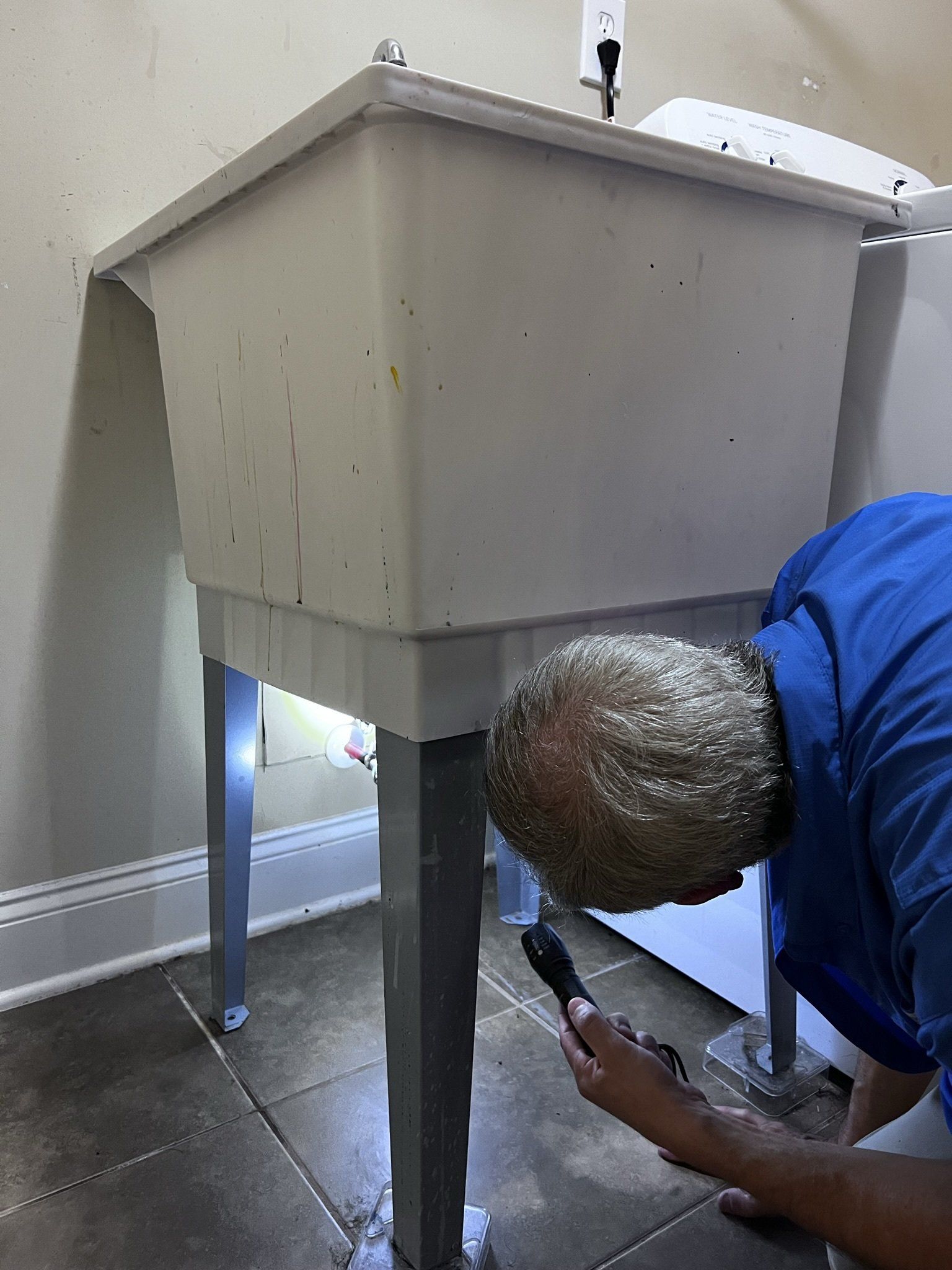 A man in a blue shirt is looking under a laundry sink.
