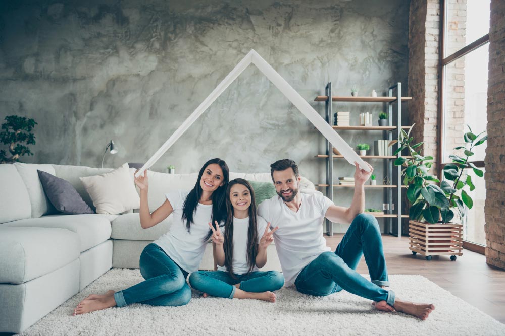 A family is sitting on the floor in a living room holding a picture of a house.