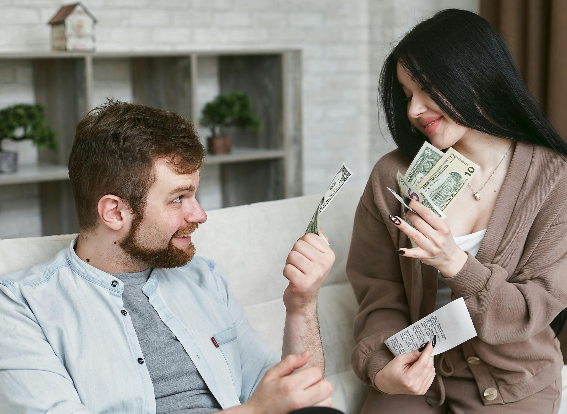 Man and woman on couch, holding cash. Man smiles while the woman counts. Indoor setting.