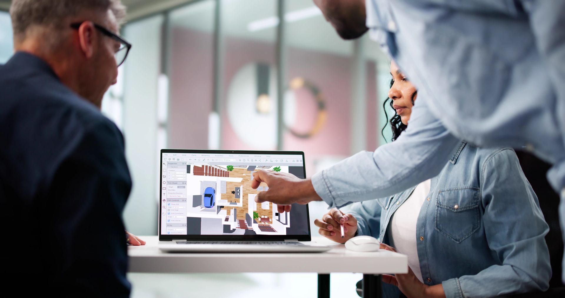 Three real estate agents reviewing floor plans on a laptop in an office setting; one points.