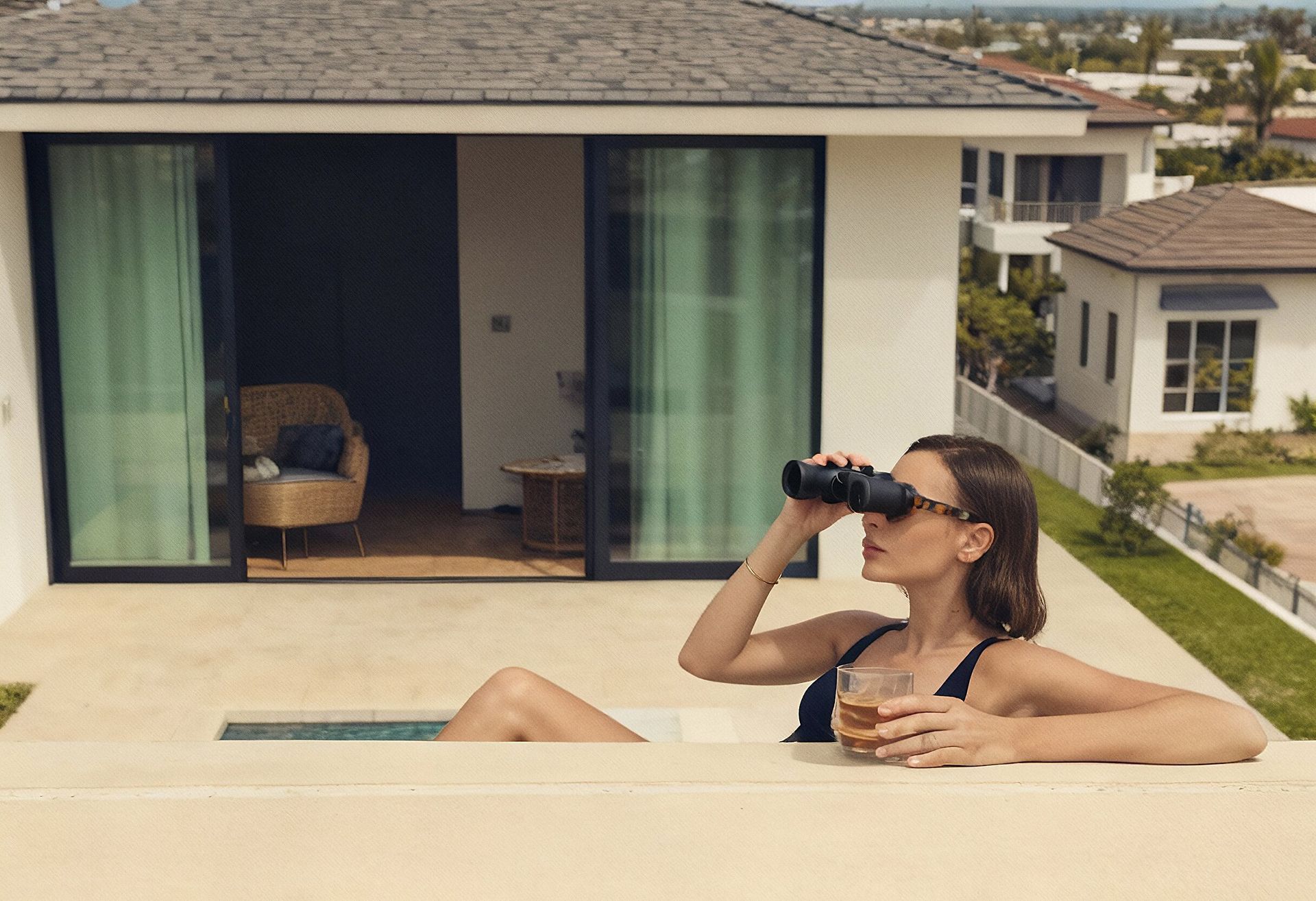 Woman using binoculars from a pool, holding a drink, with a house and view in the background.