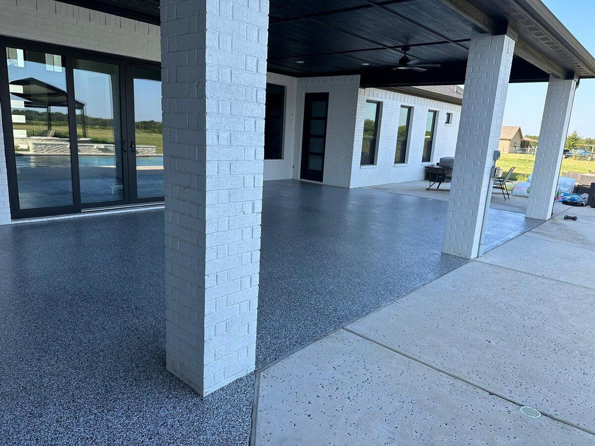Covered patio with speckled gray flooring, white pillars, and glass doors to the left.