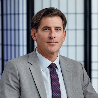 A person wearing a grey suit, light blue dress shirt, and a dark patterned tie, centered against a geometric backdrop.