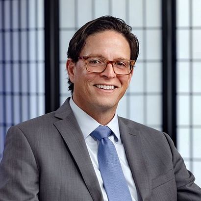 A professional headshot of a smiling person wearing glasses, a grey suit, and a blue tie against a paneled background.